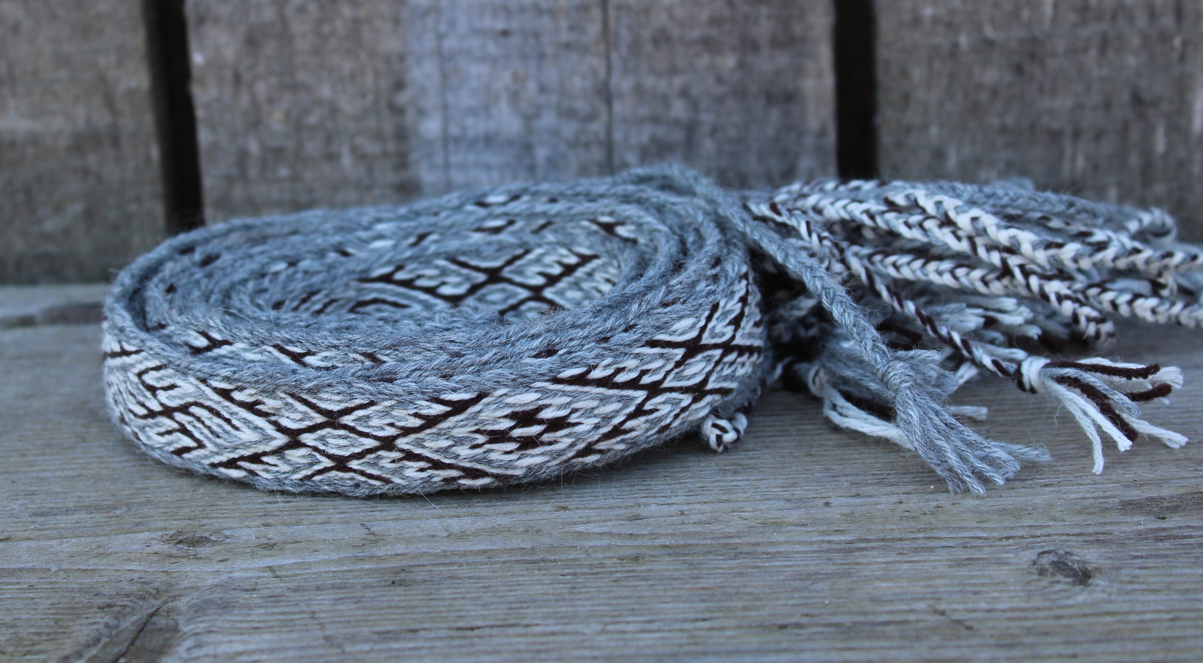 A gray and black woven rope or lariat-style strap with braided tassels, resting on a weathered wooden surface.
