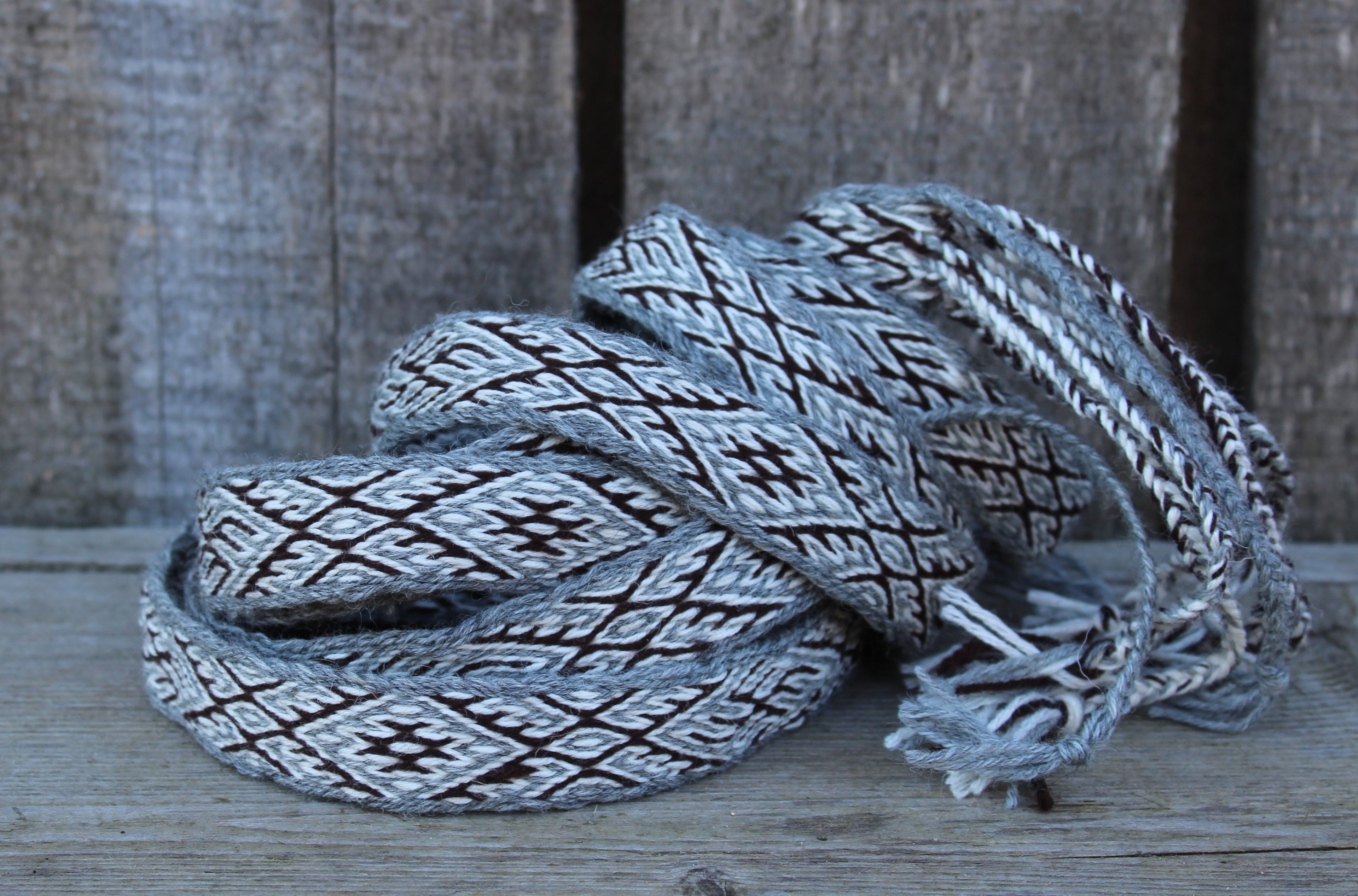 A coiled woven strap or belt with a gray, white, and black geometric pattern, resting on a rustic wooden surface against a weathered wood plank background.