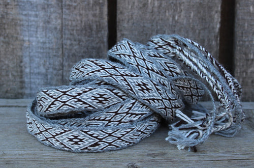 A coiled woven strap or belt with a gray, white, and black geometric pattern, resting on a rustic wooden surface against a weathered wood plank background.