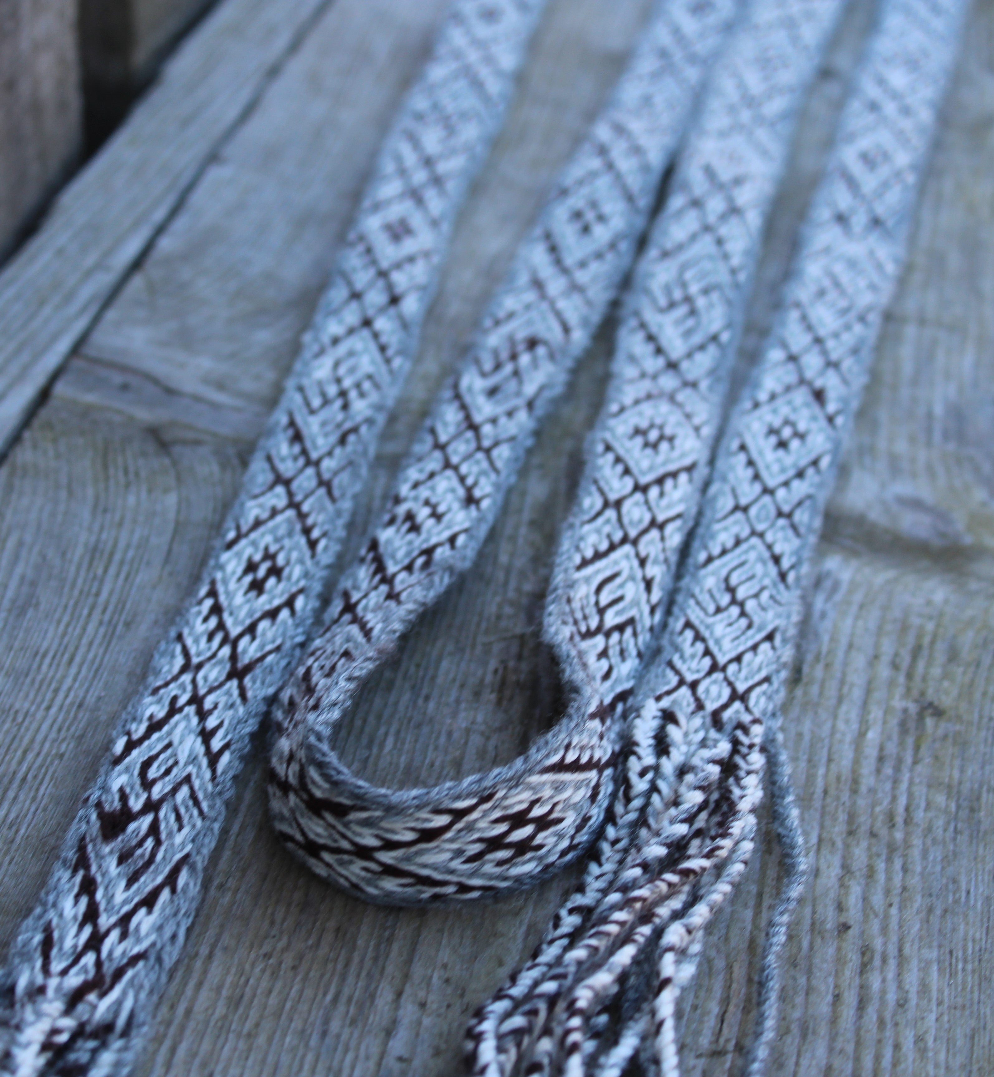 A close-up of a woven textile strap with a gray and white geometric pattern and fringed ends, laid diagonally on weathered wooden planks.