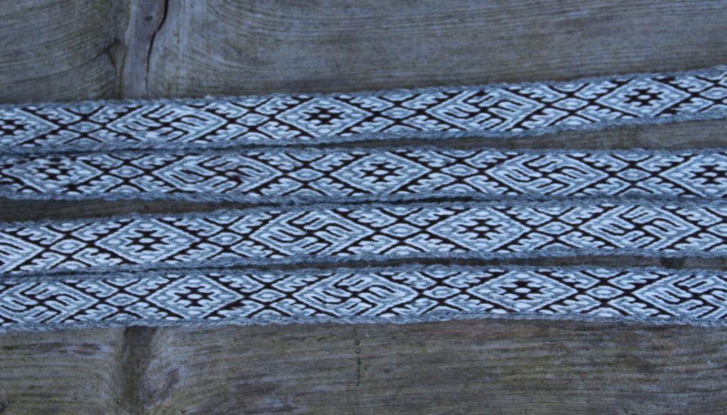 A close-up of a woven textile or strap with a repeating geometric diamond pattern in black and off-white, laid horizontally across a rustic wooden surface.