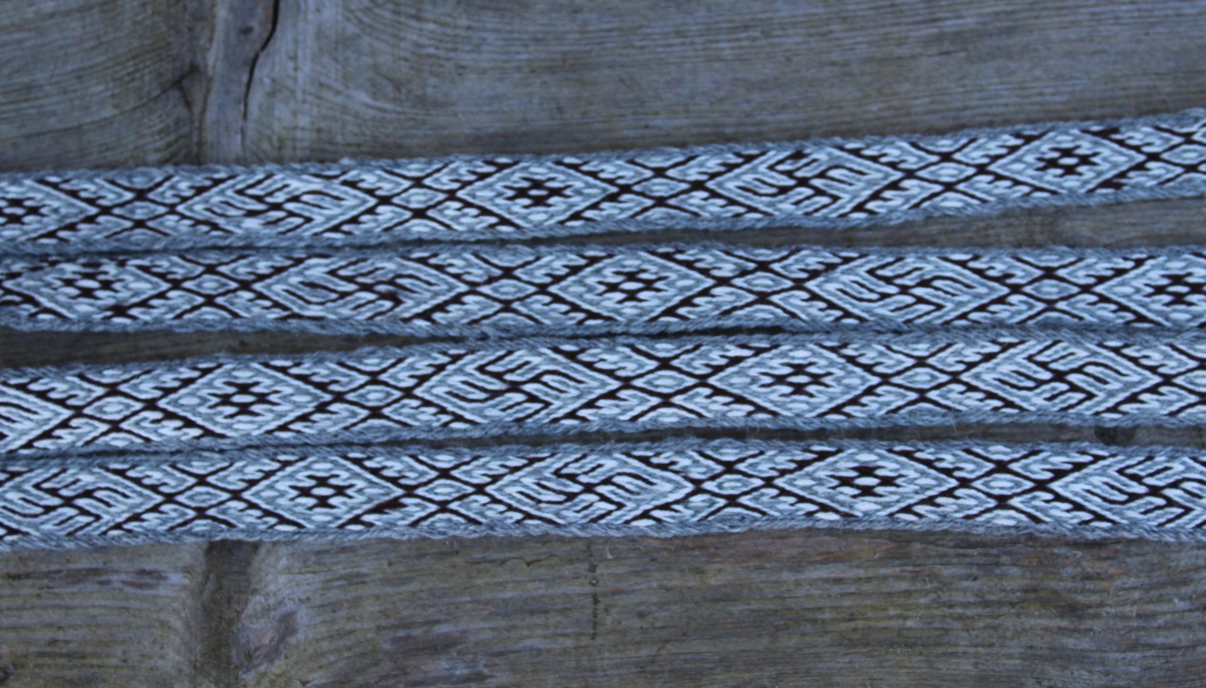 A close-up of a woven textile or strap with a repeating geometric diamond pattern in black and off-white, laid horizontally across a rustic wooden surface.