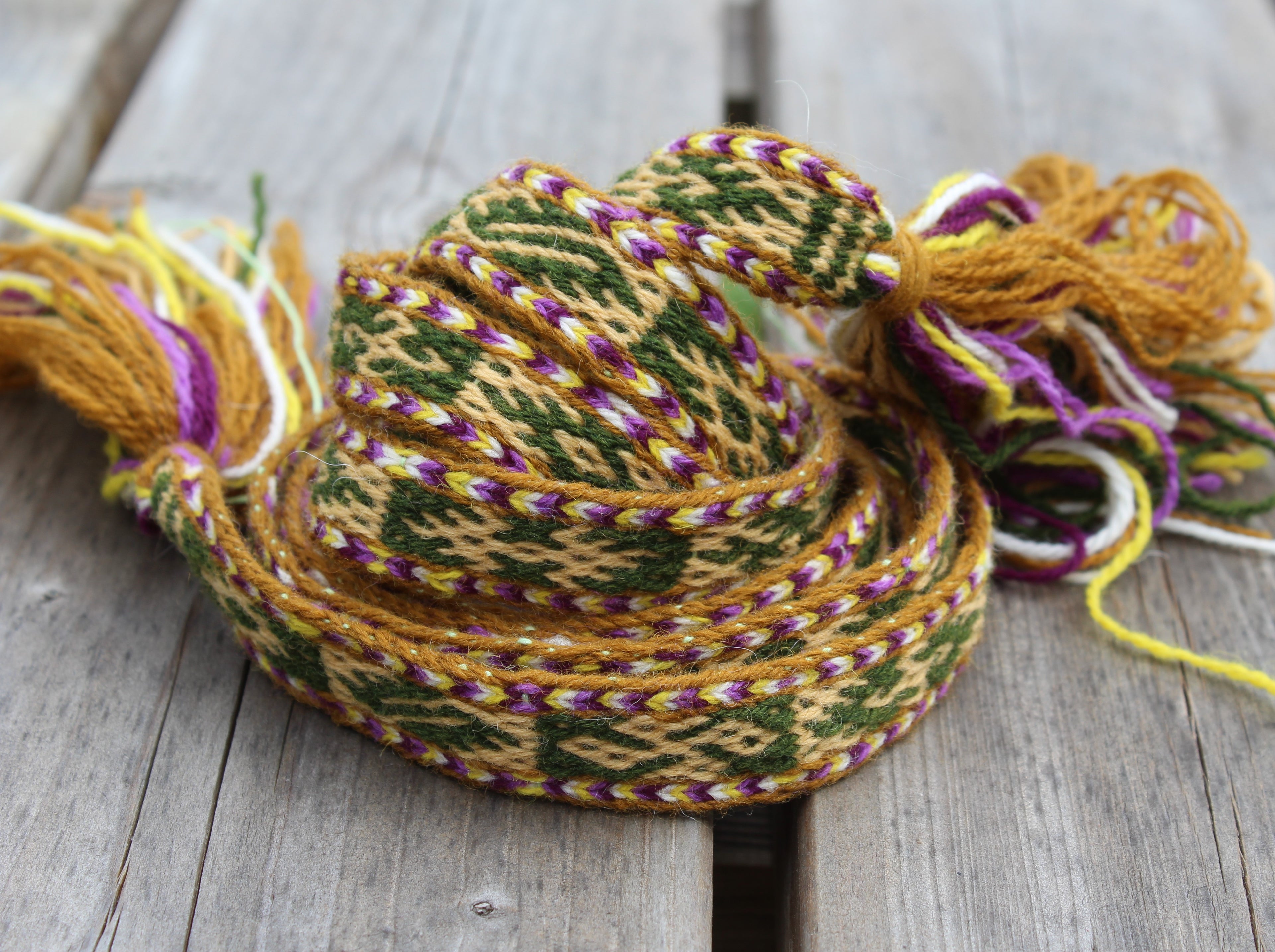 A coiled multicolor woven textile with tassels, featuring a geometric pattern in mustard yellow, green, purple, and white, displayed on a weathered gray wooden surface.