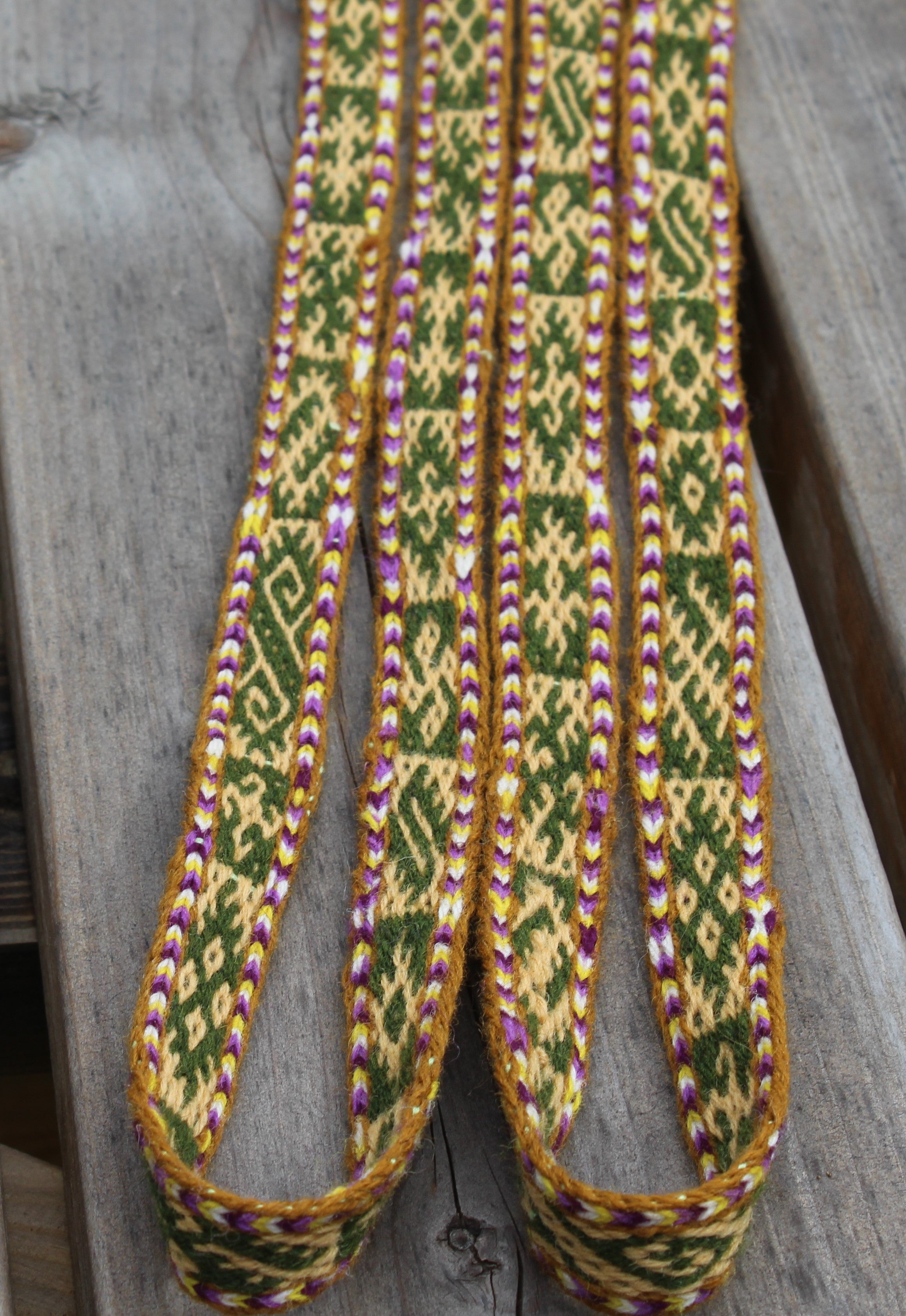 A close-up of a woven strap or belt with a geometric pattern in green, beige, and purple, laid flat on a weathered gray wooden surface.