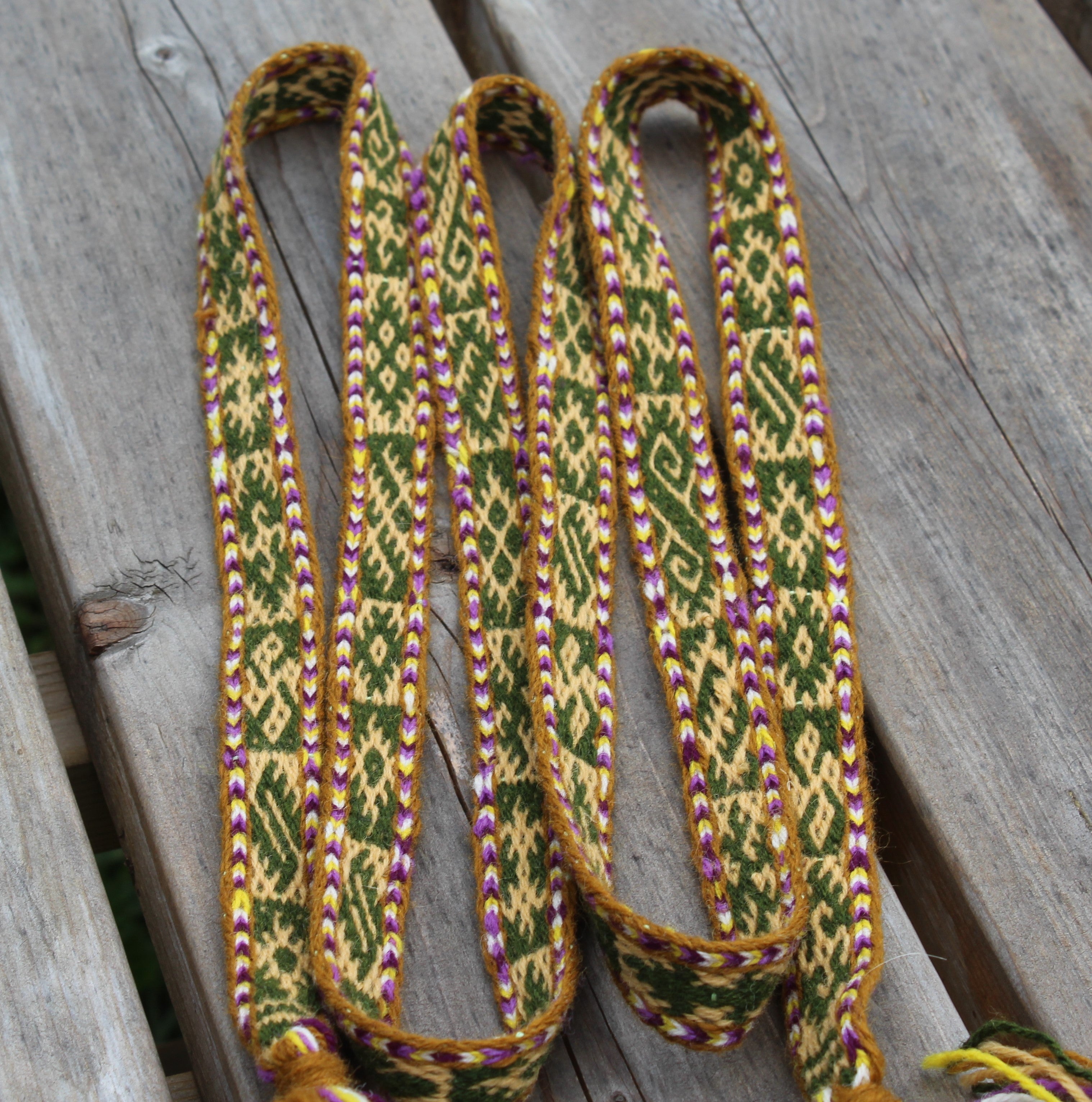 A close-up of a woven strap or belt with a geometric pattern in green, yellow, and purple, laid out on weathered gray wooden planks.