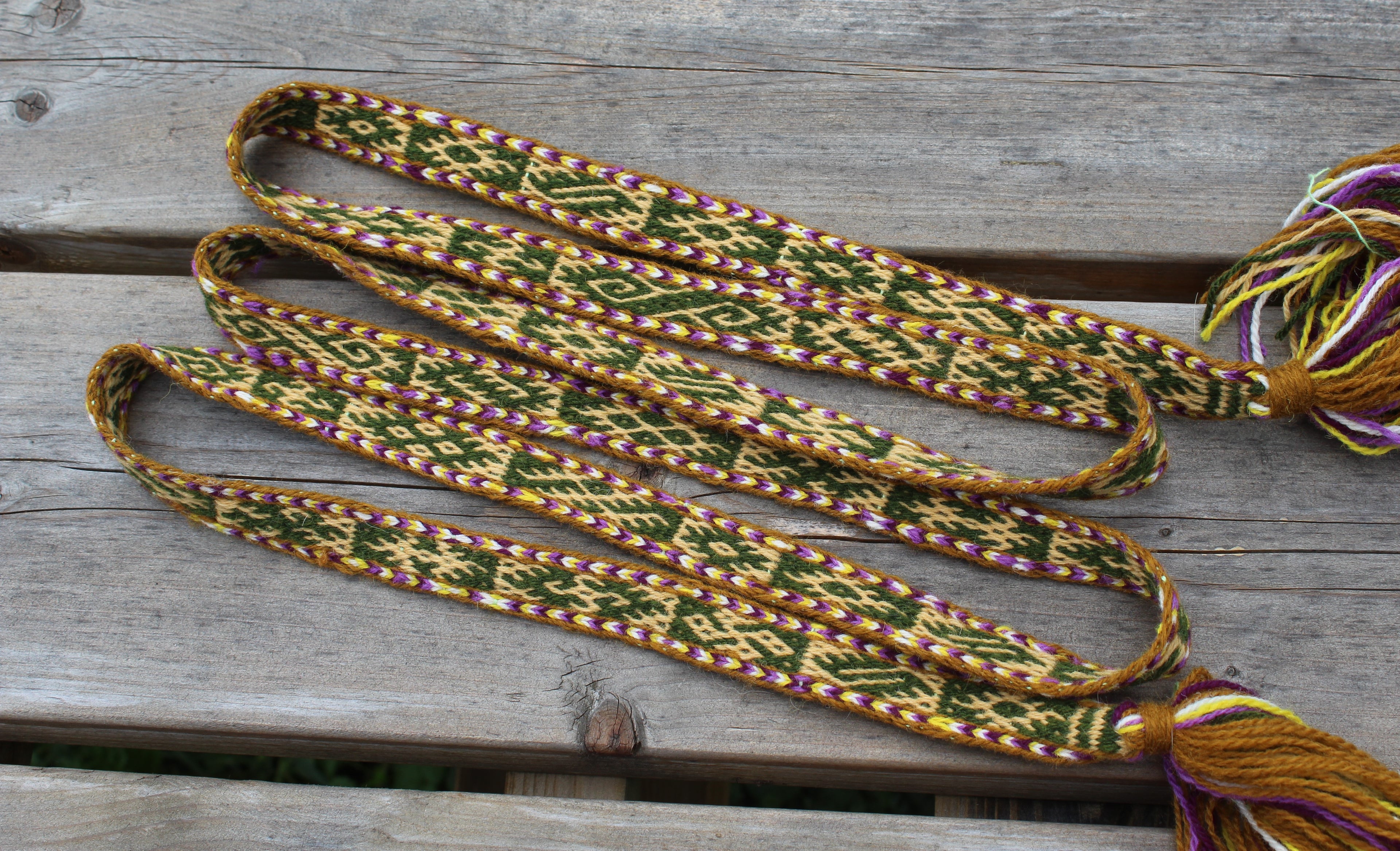 A handmade braided friendship cord or lanyard with geometric patterns in green, gold, purple, and white, featuring tassels on both ends, laid out on a weathered gray wooden surface.