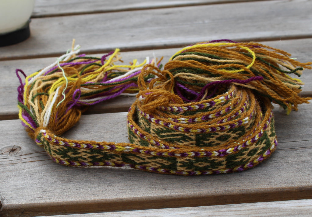 A multicolor woven textile accessory, likely a belt or strap, with braided patterns in yellow, green, purple, and white, featuring frayed tassels, displayed on a weathered wooden surface.