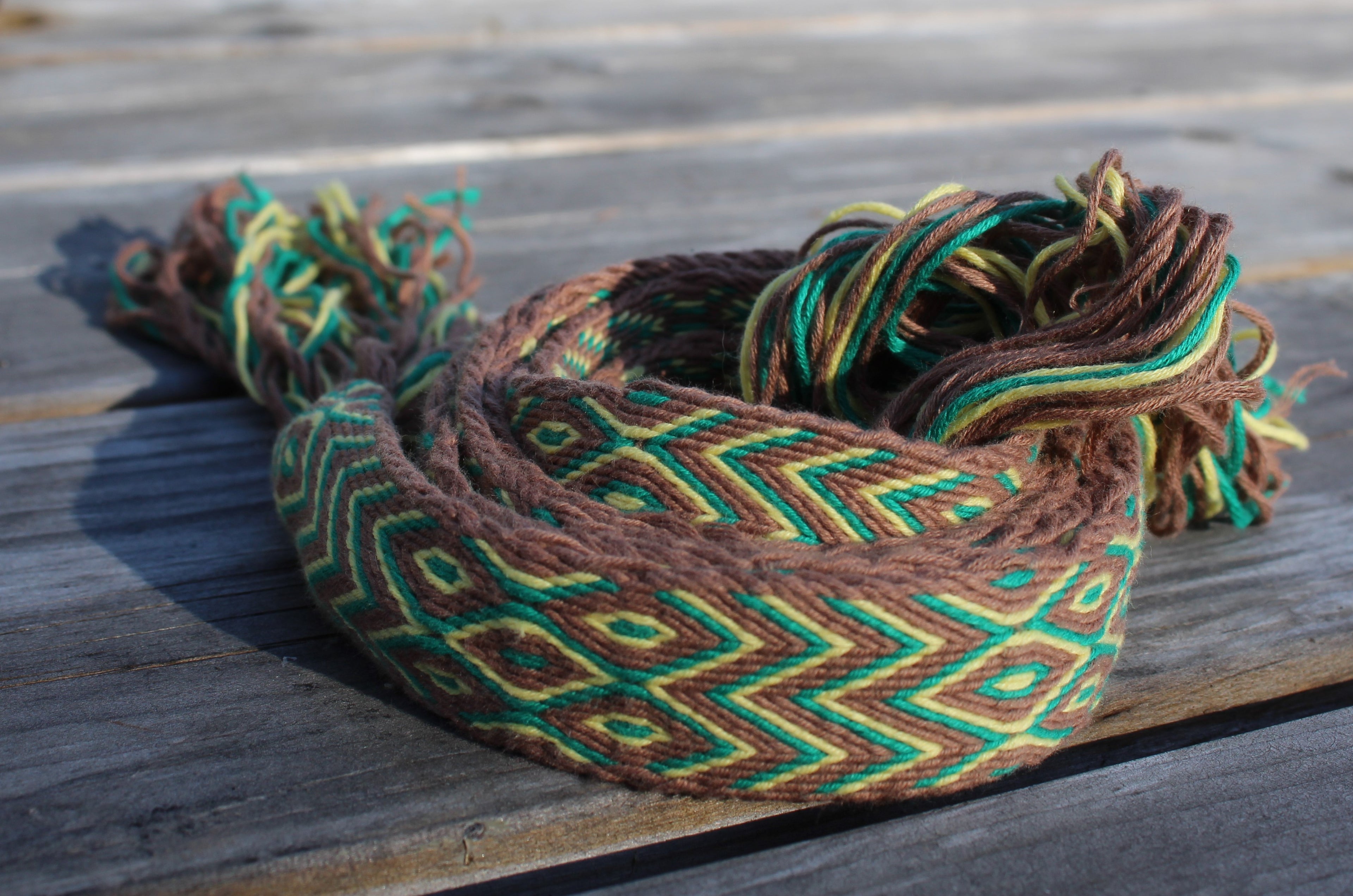 A woven rope accessory, likely a belt or wrap, featuring a geometric pattern in brown, green, and yellow, resting on a weathered wooden surface.