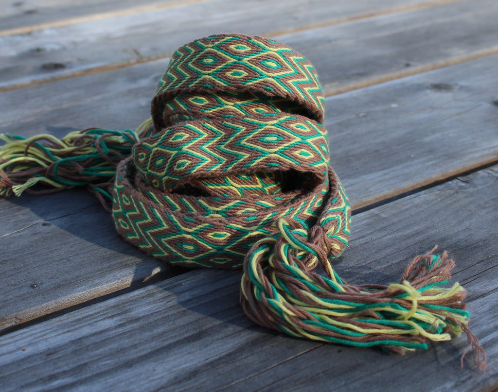 A woven multicolor scarf with a geometric pattern in green, brown, and yellow, featuring tassels, resting on weathered gray wooden planks.