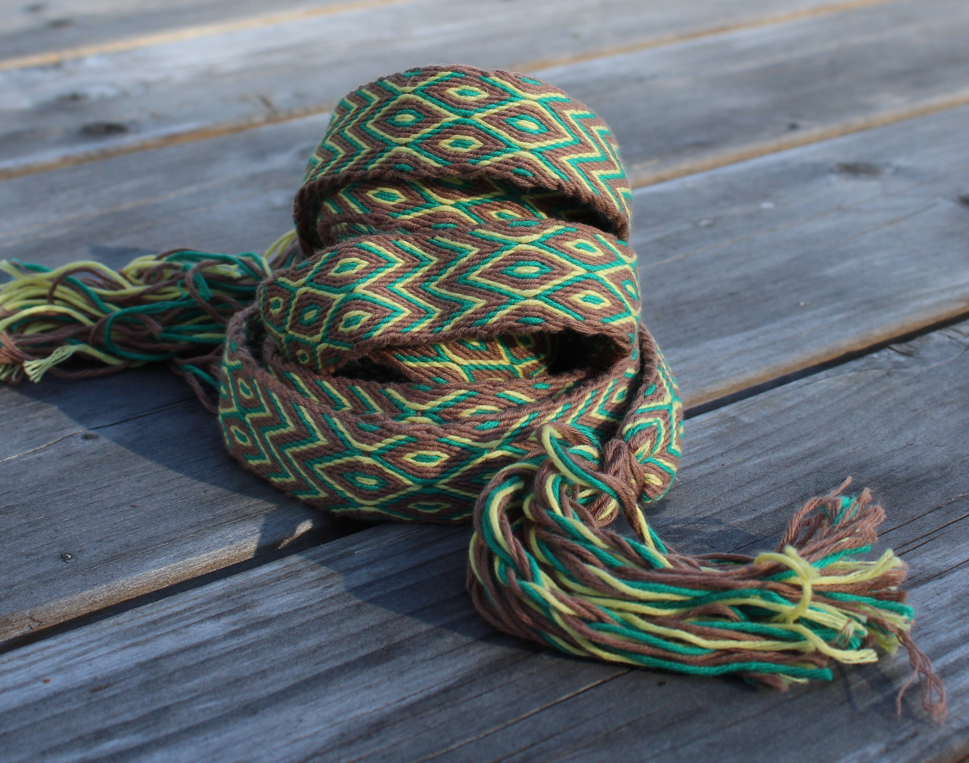 A woven multicolor scarf with a geometric pattern in green, brown, and yellow, featuring tassels, resting on weathered gray wooden planks.