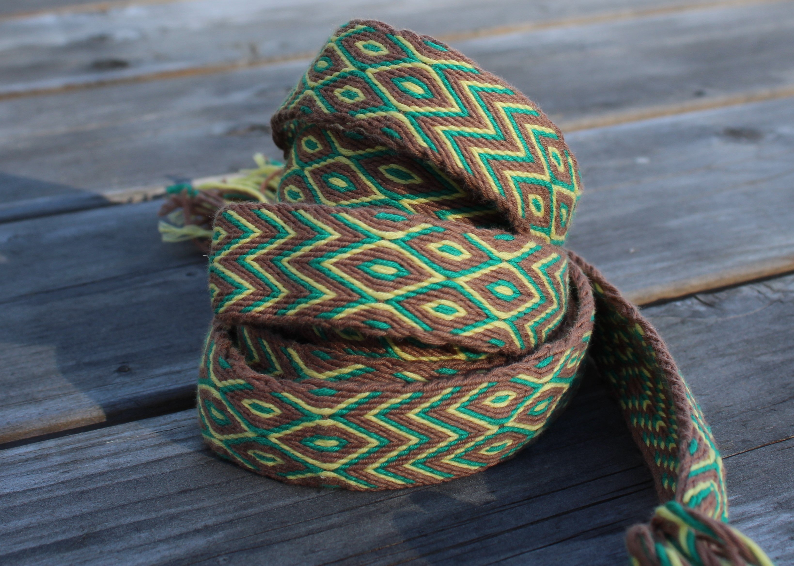 A coiled woven rope or strap with a geometric diamond pattern in green, brown, and yellow, resting on weathered wooden planks.