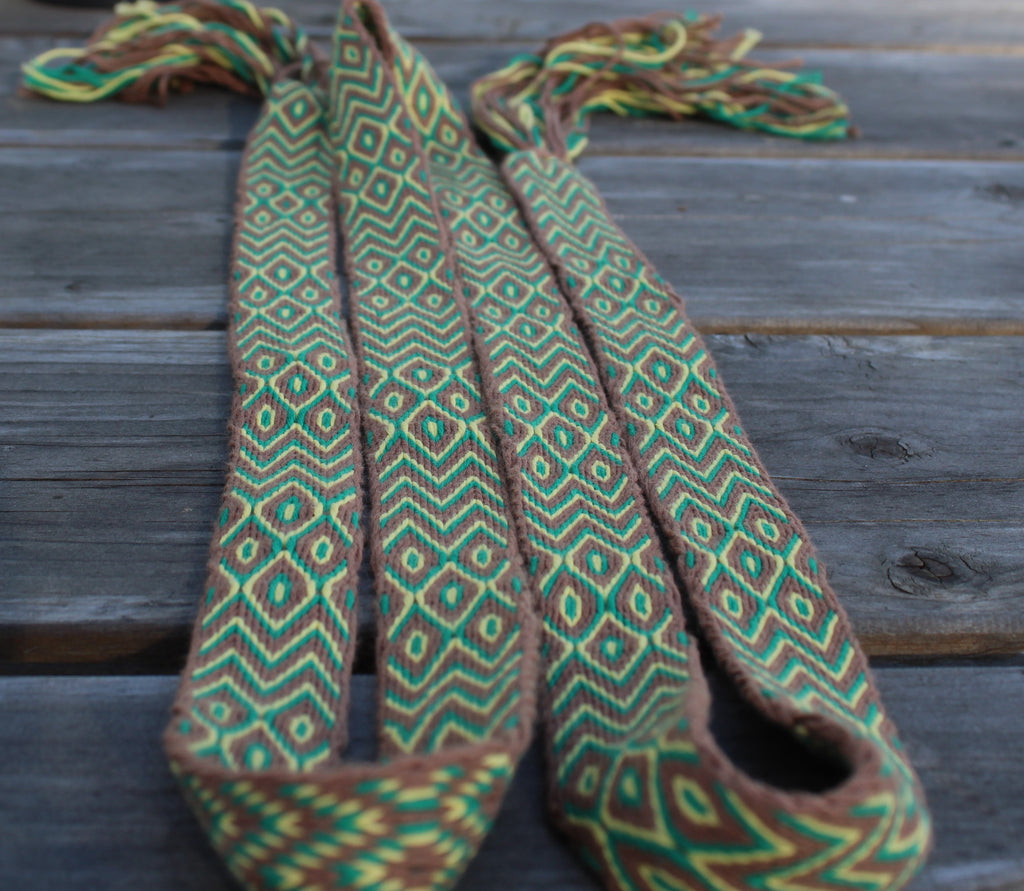 A woven textile strap or belt with a geometric diamond pattern in green, brown, and yellow, laid flat on a weathered gray wooden surface.