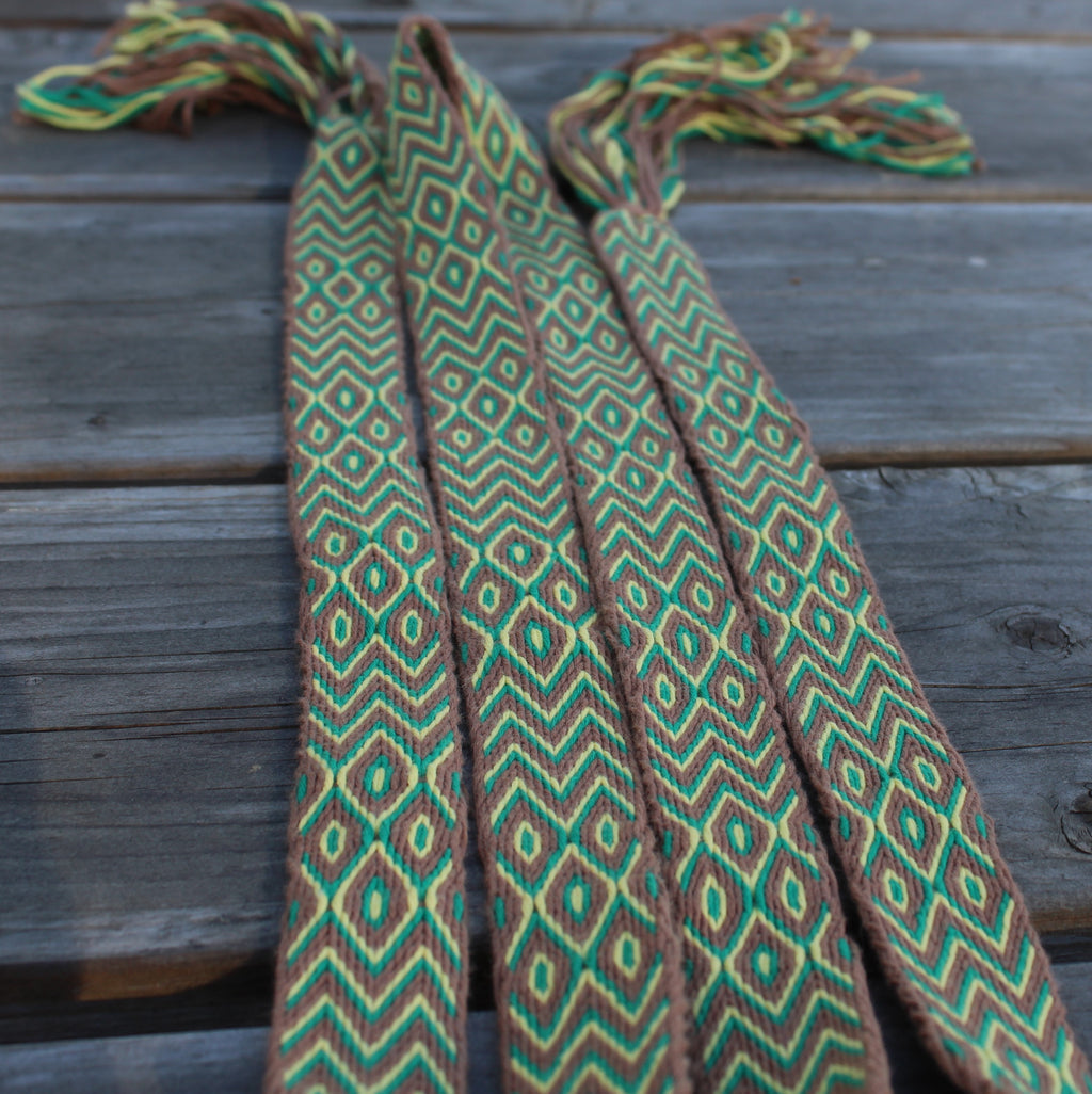 A woven waist wrap or scarf with a geometric diamond pattern in green, brown, and yellow, laid flat on a weathered gray wooden plank surface.