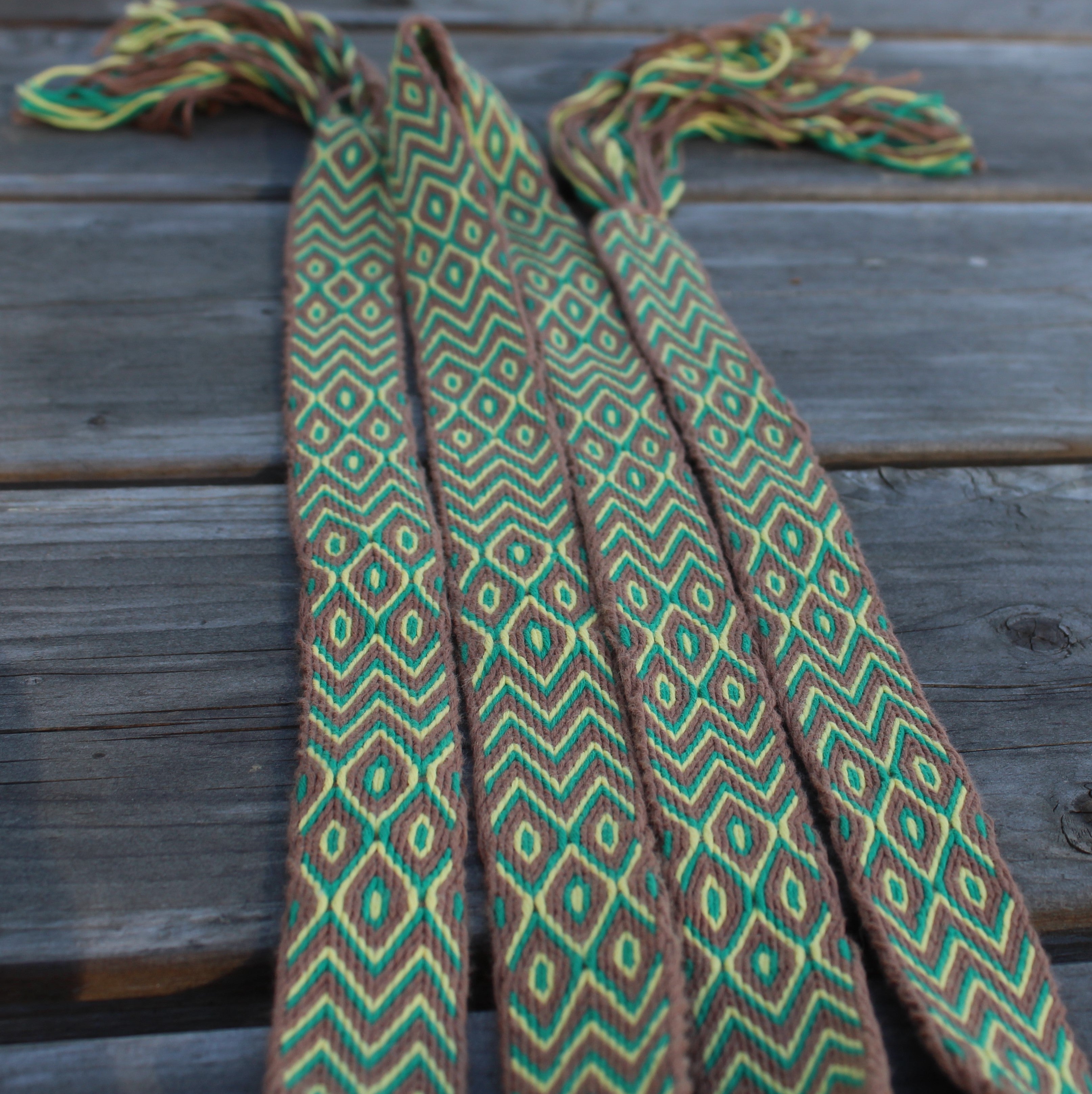 A woven waist wrap or scarf with a geometric diamond pattern in green, brown, and yellow, laid flat on a weathered gray wooden plank surface.