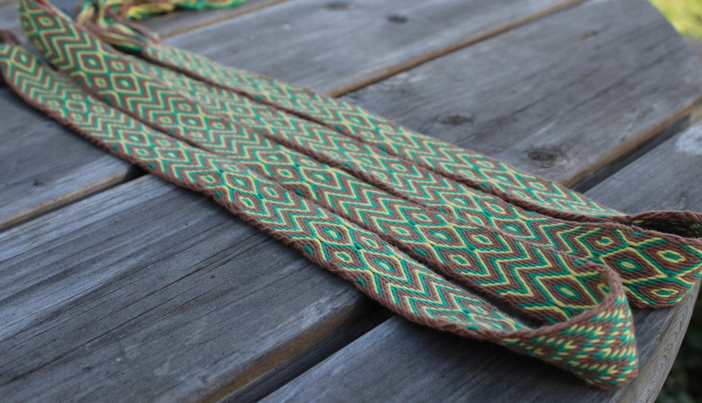 A woven textile strap with a geometric pattern in green, brown, and yellow, laid diagonally across weathered gray wooden planks.