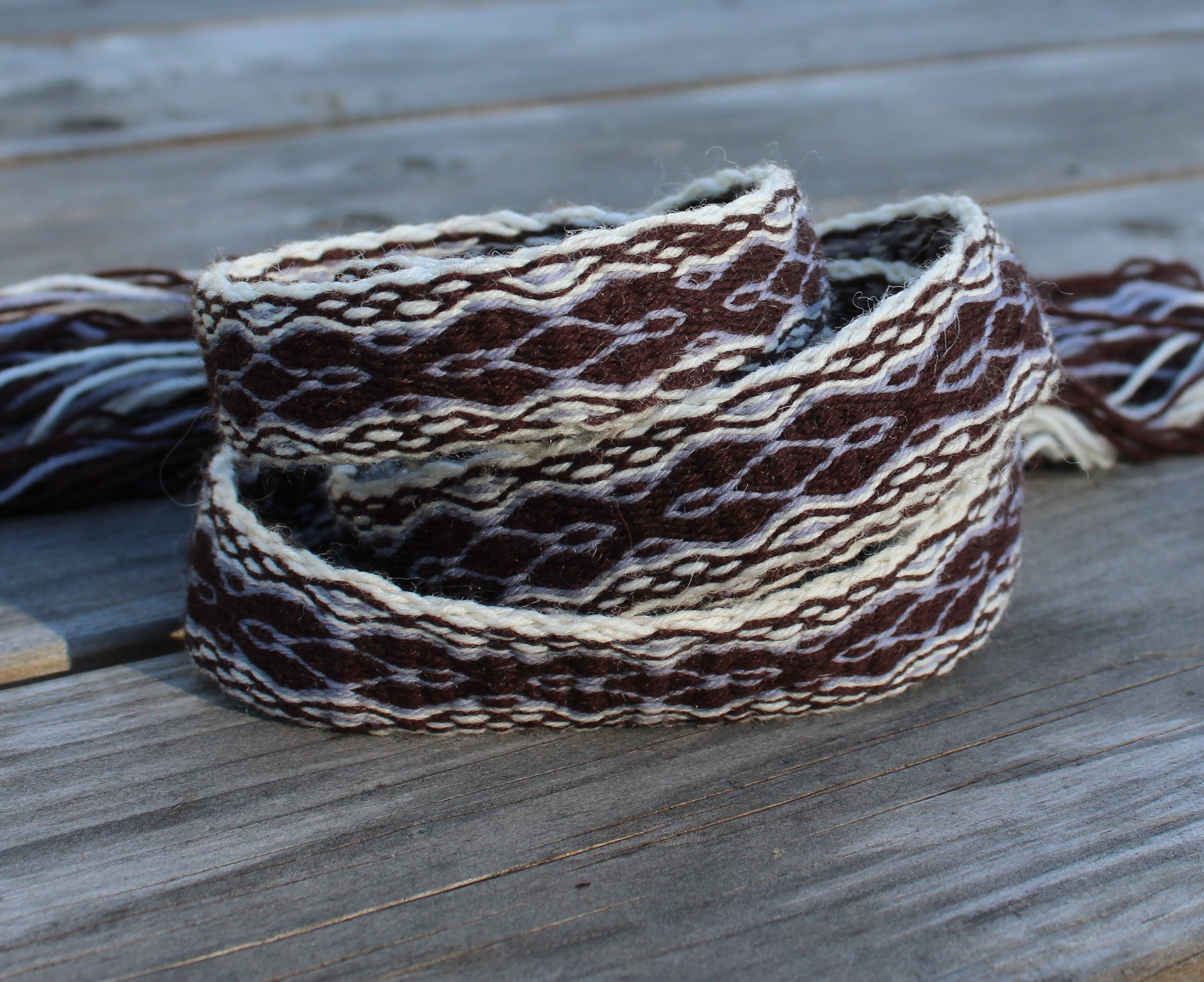 A woven wool belt with a brown, cream, and white geometric pattern, coiled on a weathered gray wooden surface.