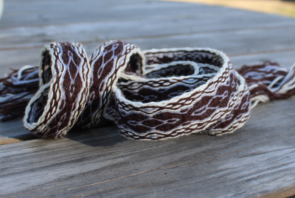 A close-up of a woven textile accessory, likely a belt or strap, featuring a brown and cream geometric pattern, resting on a weathered gray wooden surface.