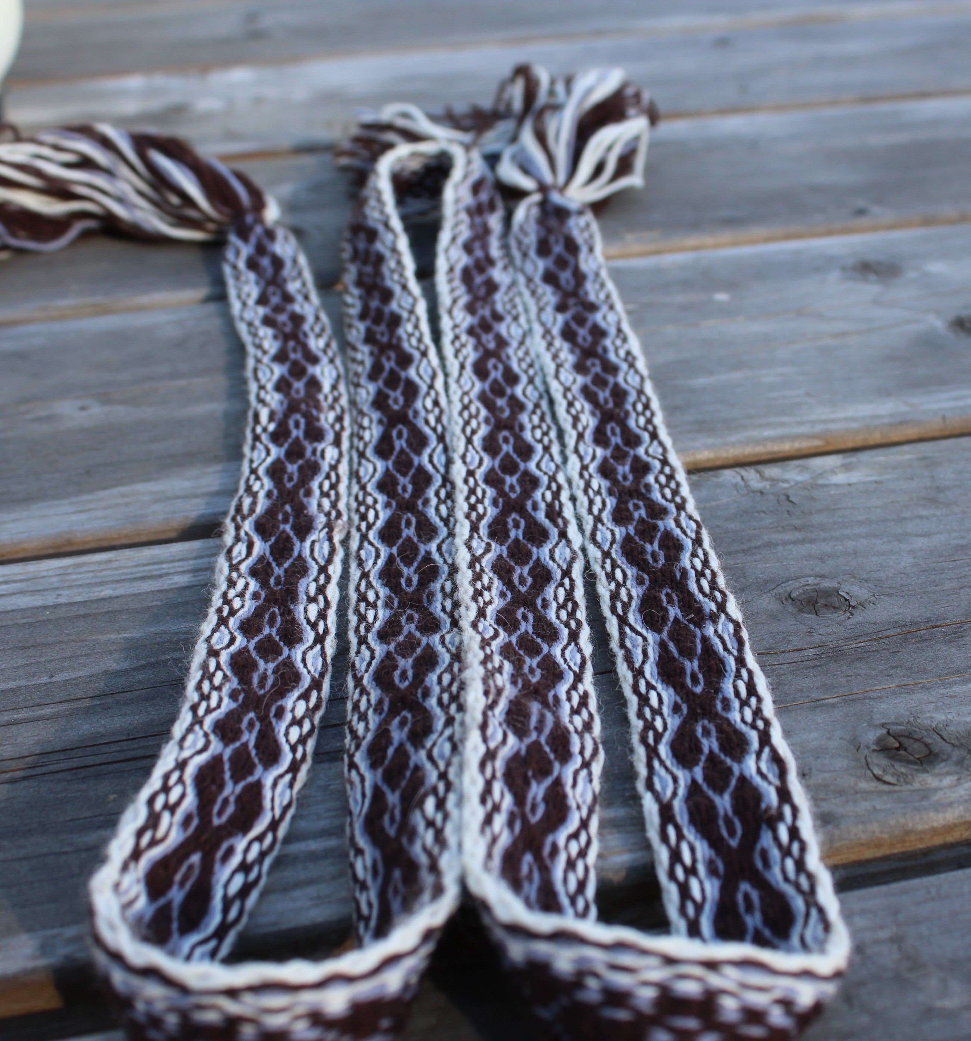 Three long braided rope straps with tassel ends, laid flat on a weathered gray wooden surface. The braided pattern is a mix of dark brown, cream, and light gray.