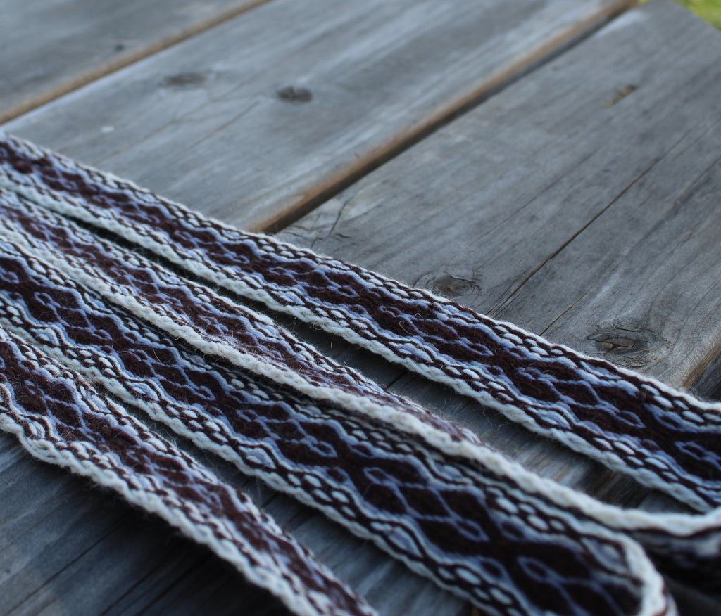 A close-up of a braided woven strap with a geometric pattern in dark brown, cream, and gray, laid diagonally across weathered gray wooden planks.