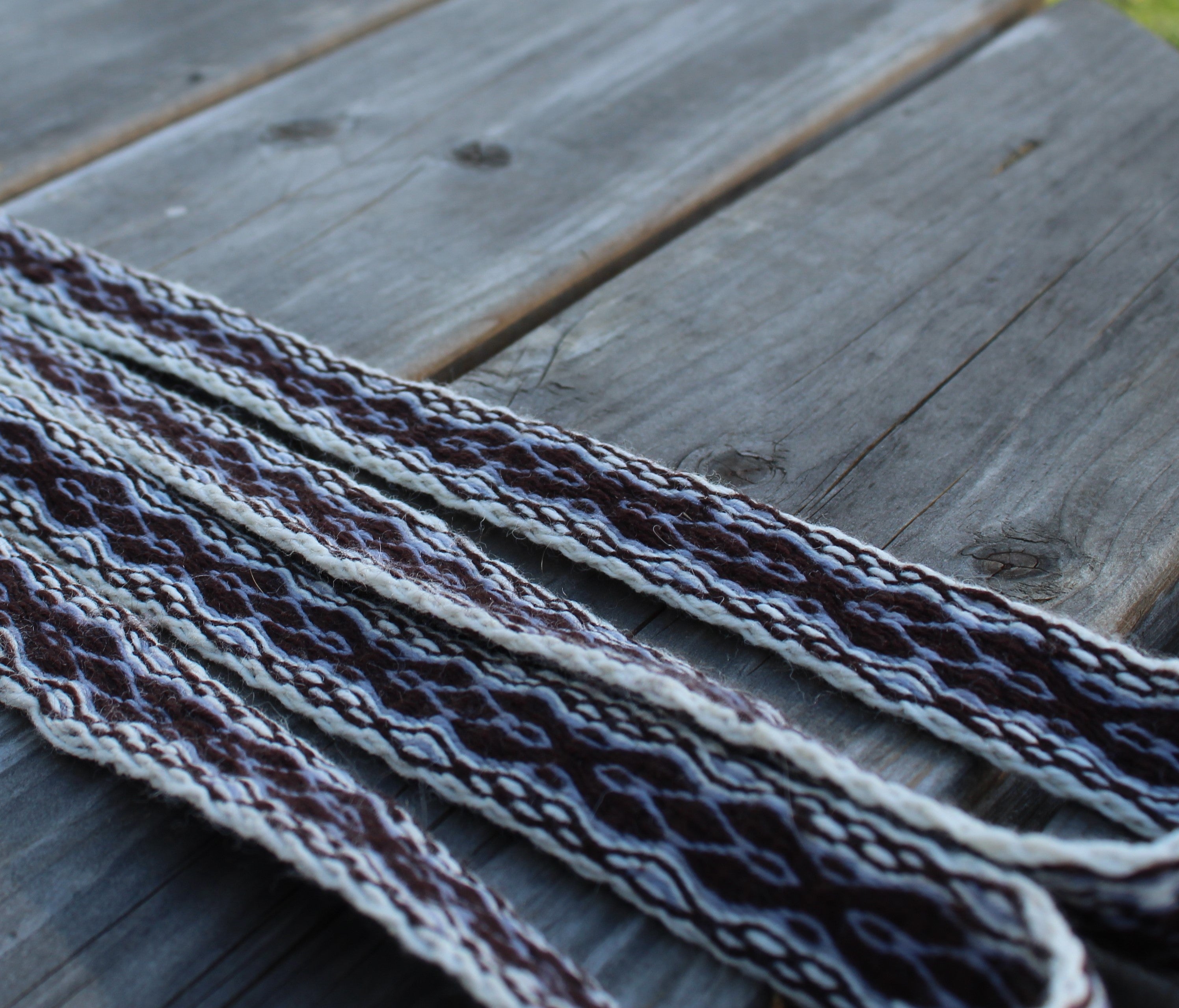 A close-up of a braided woven strap with a geometric pattern in dark brown, cream, and gray, laid diagonally across weathered gray wooden planks.