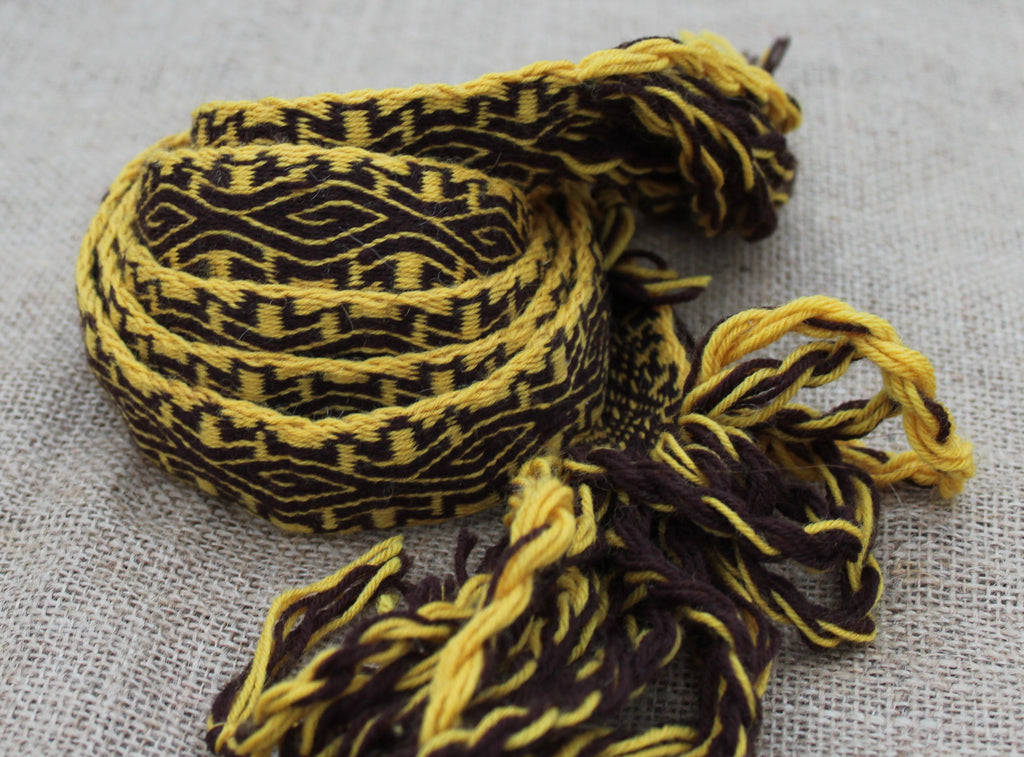 A woven braided cord or strap with a geometric pattern in yellow and dark brown, finished with frayed tassels, laid on a textured light gray fabric background.
