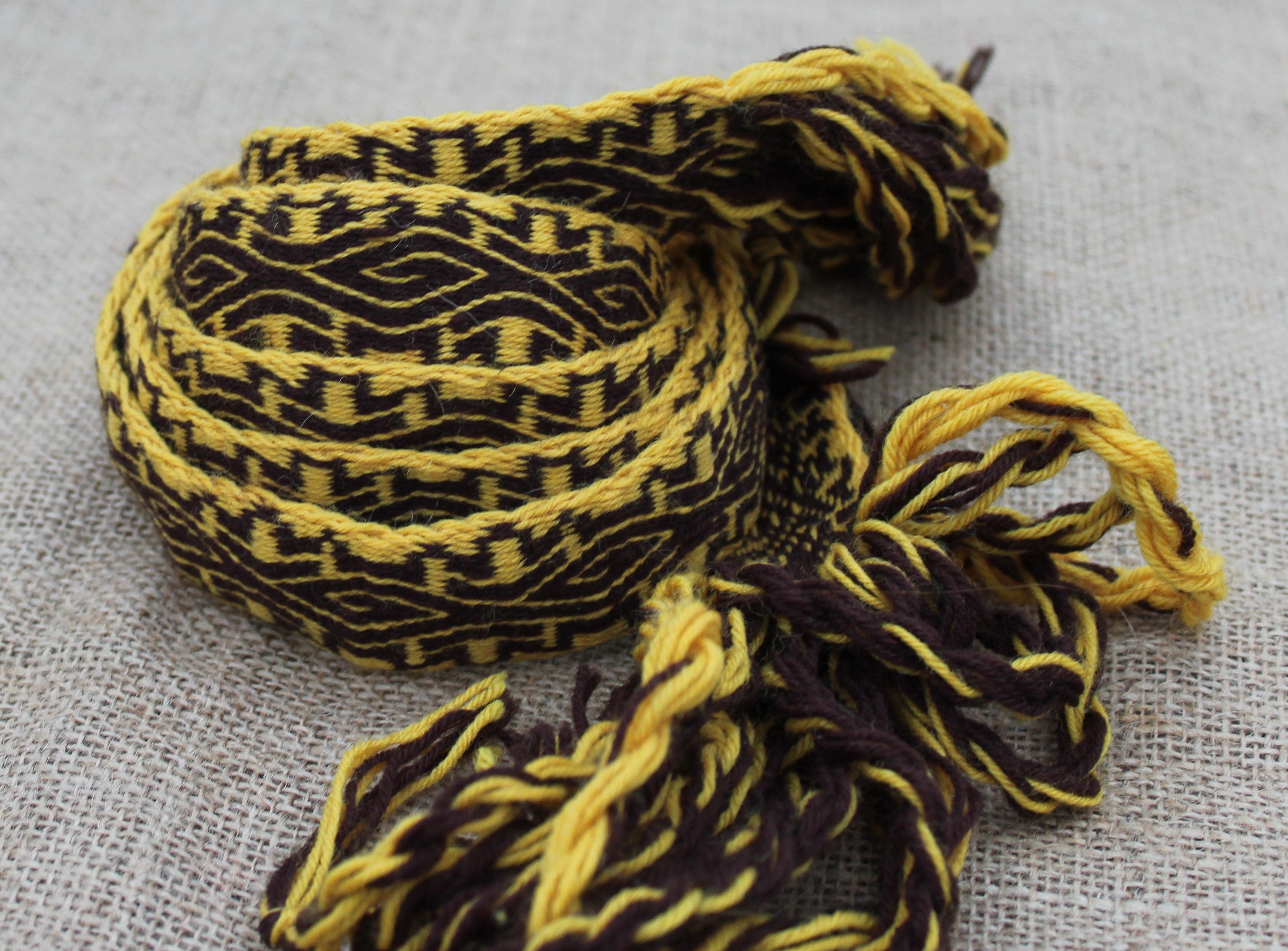 A woven braided cord or strap with a geometric pattern in yellow and dark brown, finished with frayed tassels, laid on a textured light gray fabric background.