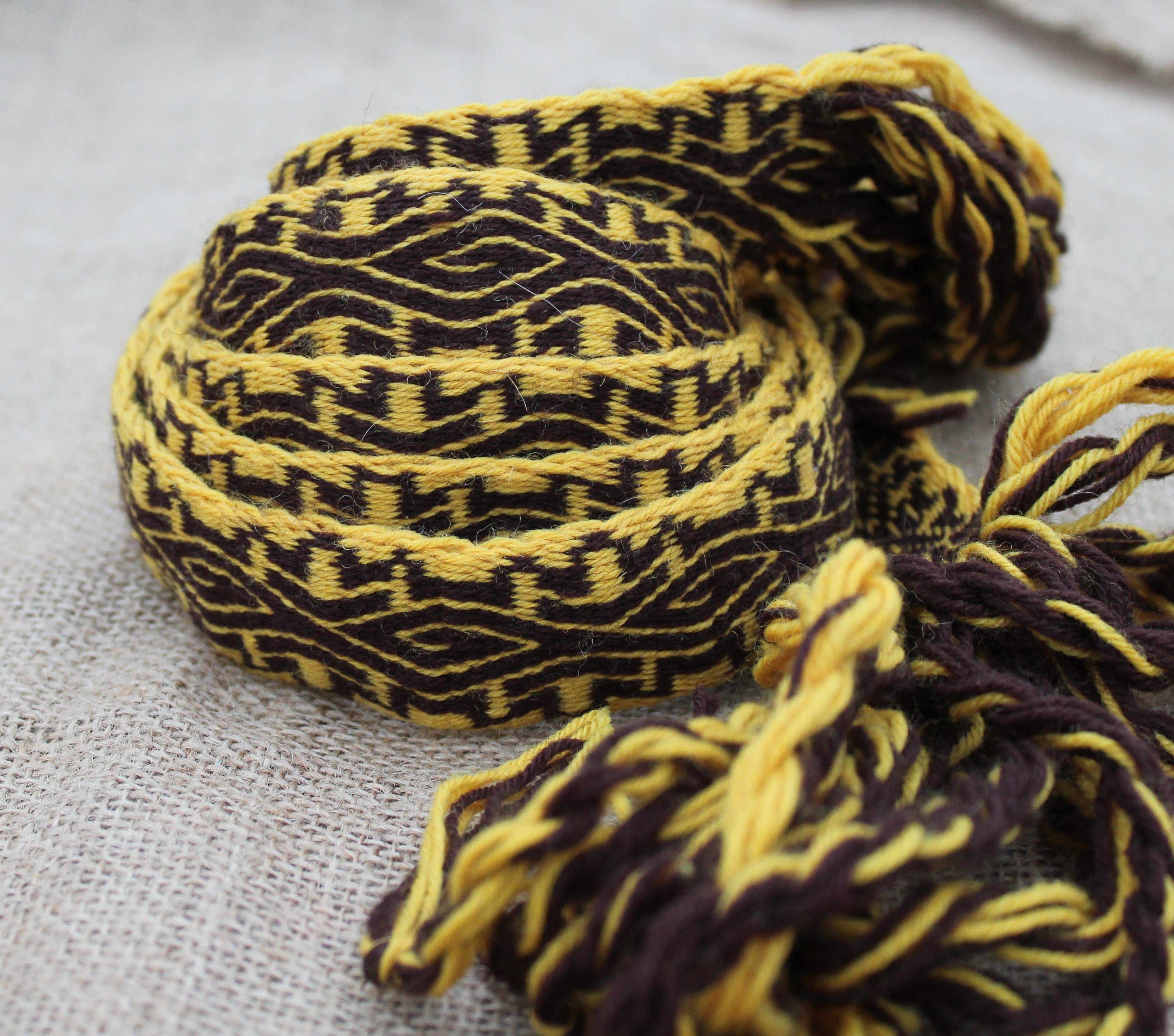 A close-up of a woven textile strap or belt with a geometric pattern in dark brown and mustard yellow, featuring frayed tassels, resting on a light gray linen surface.