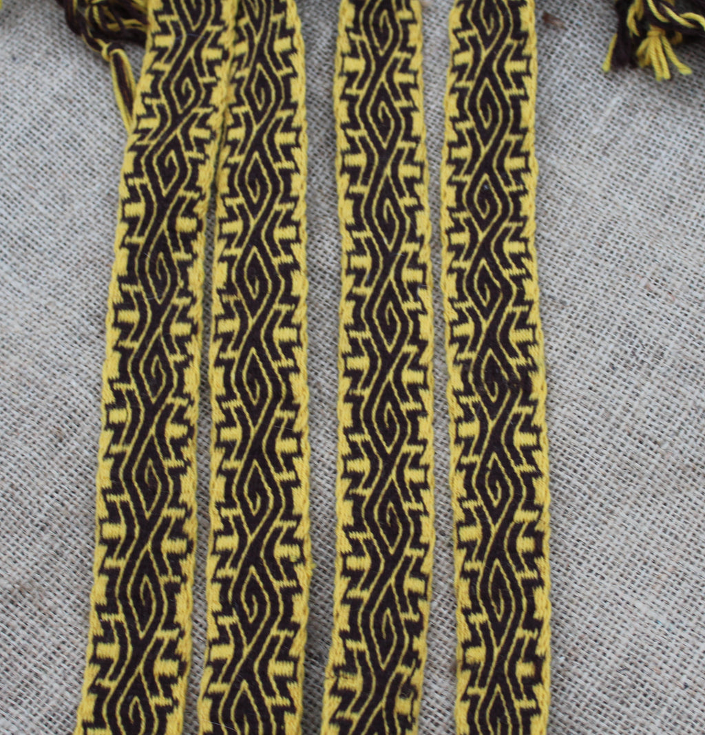 Four narrow woven straps with a bold black geometric pattern on a mustard yellow background, laid flat on a textured gray fabric surface.