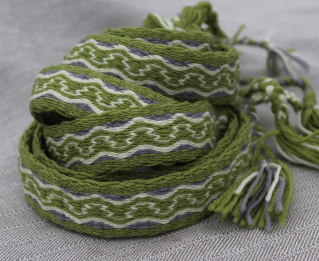 A coiled woven scarf with a geometric pattern in olive green, white, and gray, featuring knotted tassels on one end, set against a textured gray background.