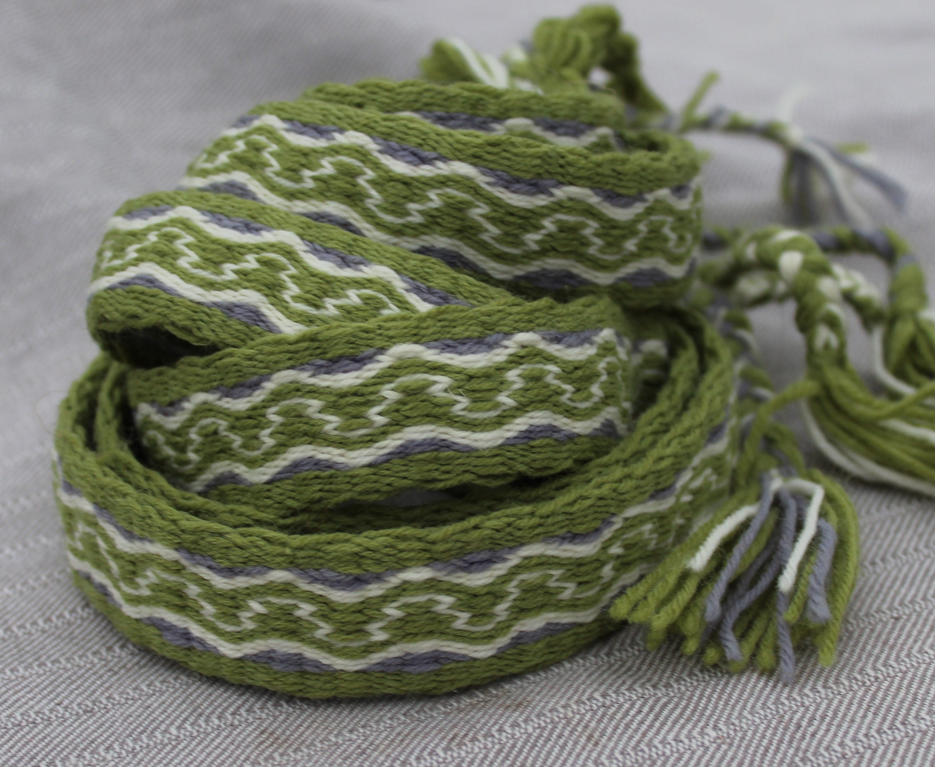 A coiled woven scarf with a geometric pattern in olive green, white, and gray, featuring knotted tassels on one end, set against a textured gray background.