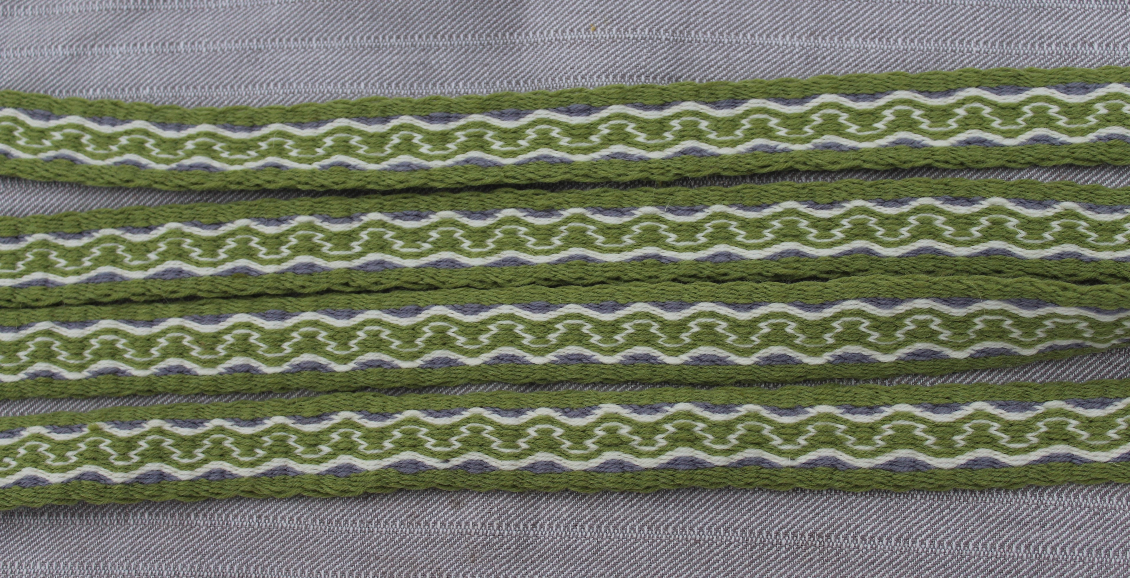 Close-up of olive green patterned ribbon or trim laid horizontally on a gray textured fabric background.