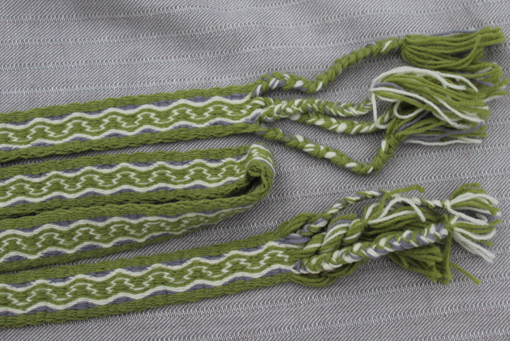 A close-up of a woven green and white striped textile accessory with braided tassel ends, laid on a textured gray fabric background.