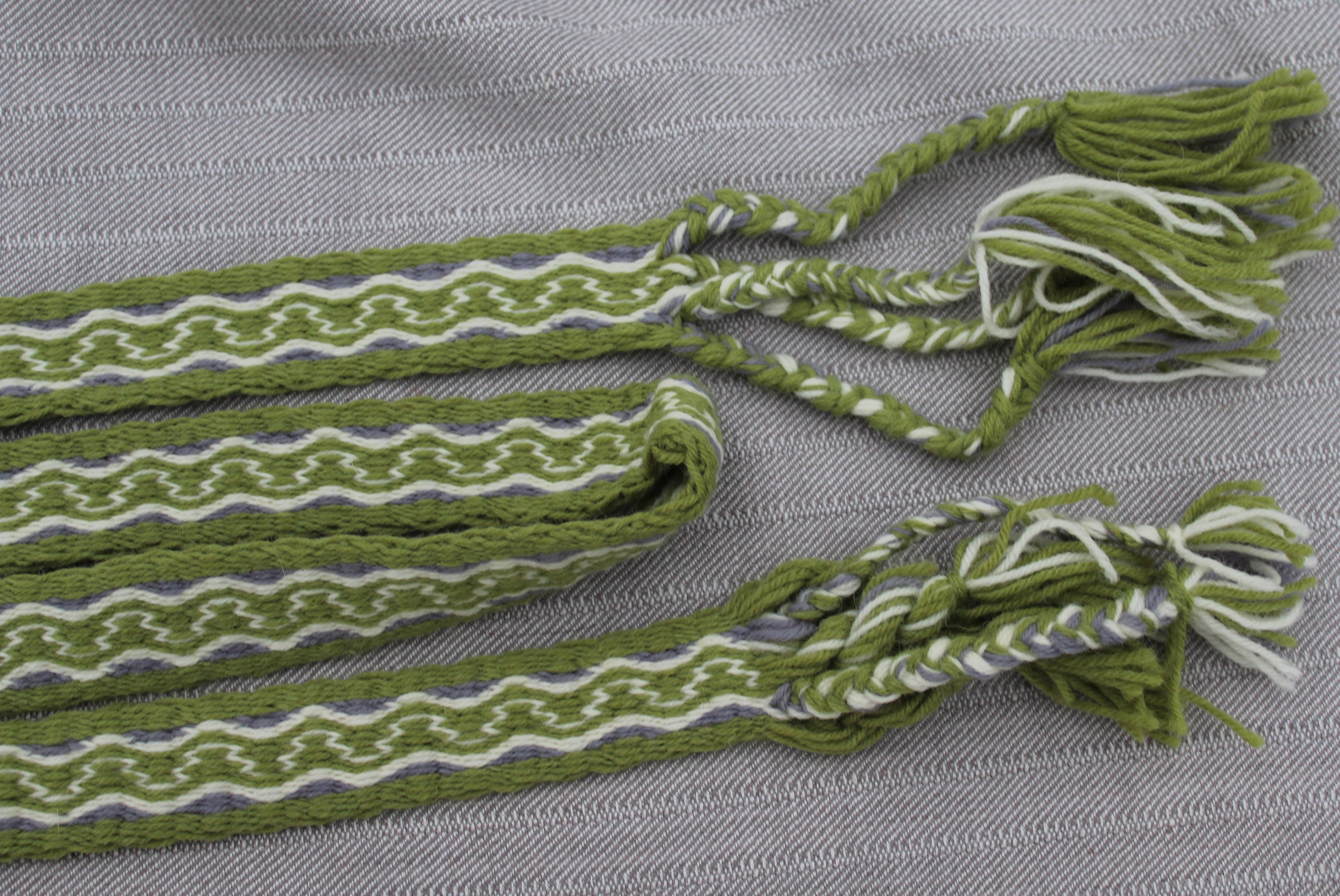 A close-up of a woven green and white striped textile accessory with braided tassel ends, laid on a textured gray fabric background.