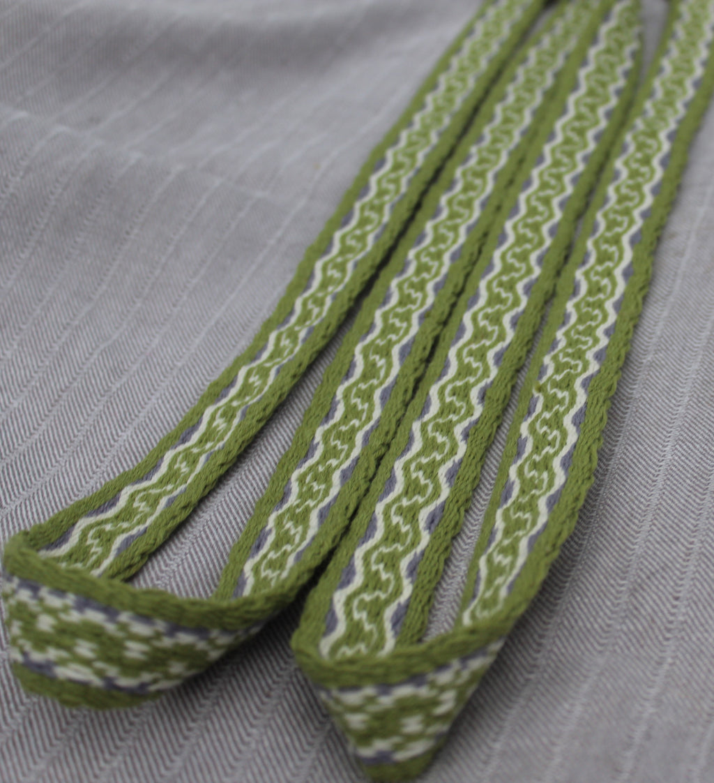 A close-up of a woven fabric strap or trim in olive green, white, and a thin blue stripe, laid diagonally on a textured light gray fabric background.