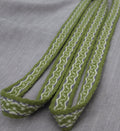A close-up of a woven fabric strap or trim in olive green, white, and a thin blue stripe, laid diagonally on a textured light gray fabric background.