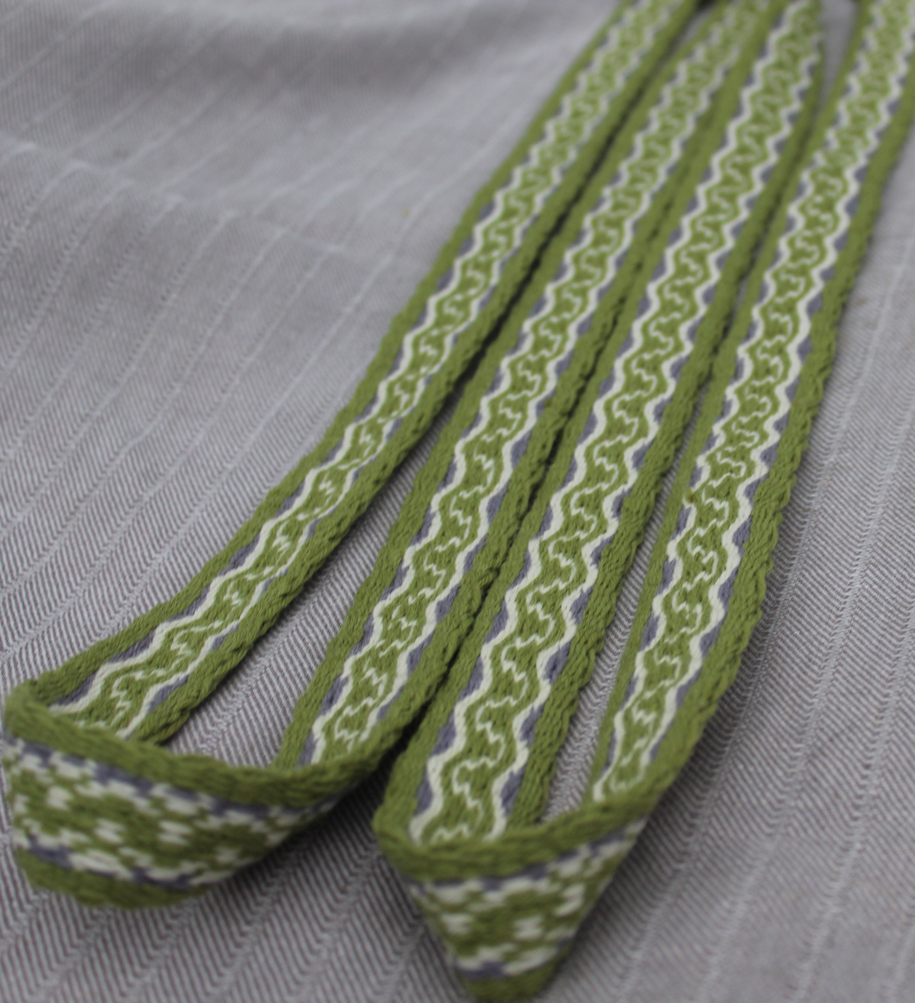 A close-up of a woven fabric strap or trim in olive green, white, and a thin blue stripe, laid diagonally on a textured light gray fabric background.