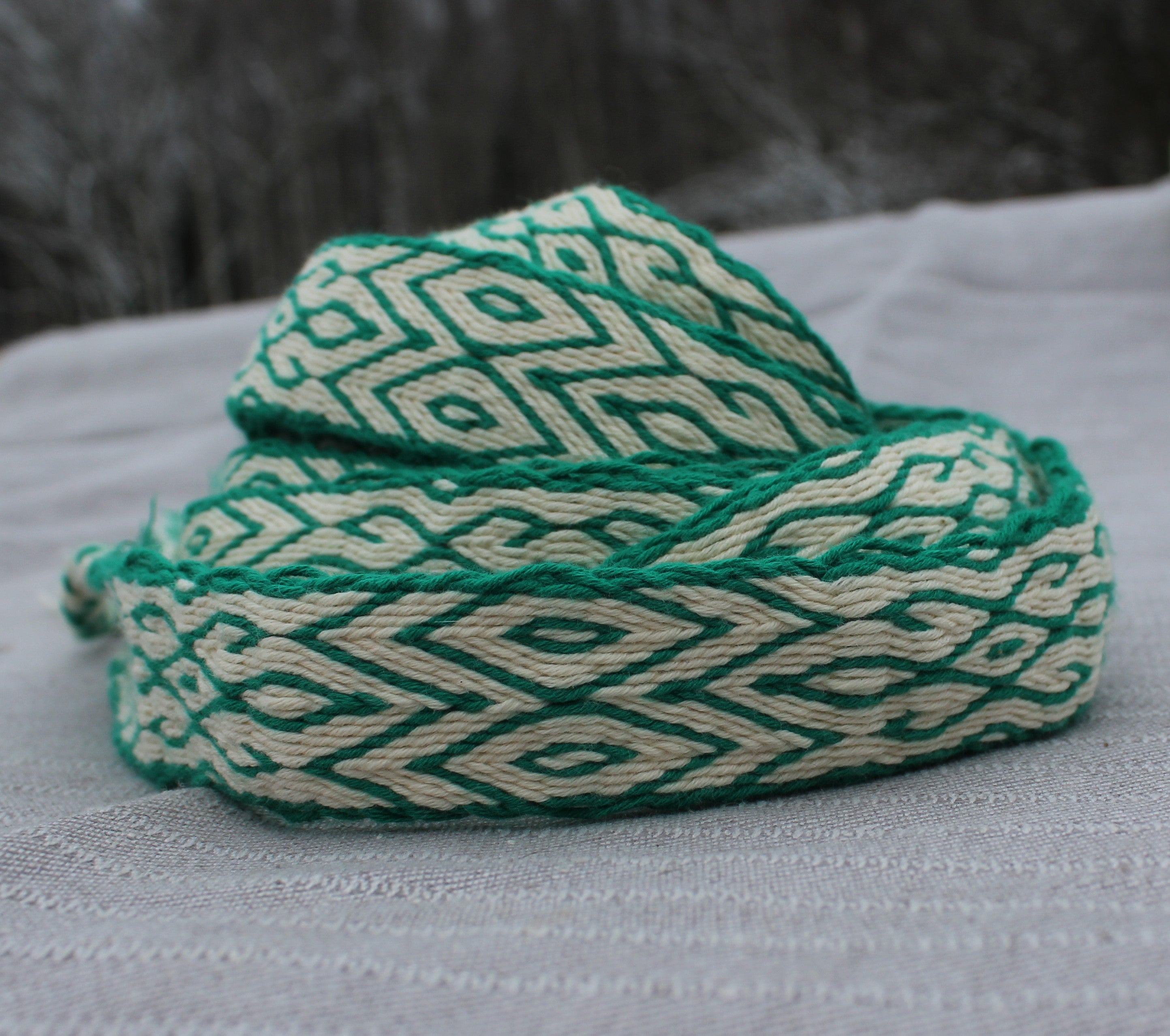 A coiled green and cream woven strap with a geometric diamond pattern, resting on a textured light gray fabric surface.