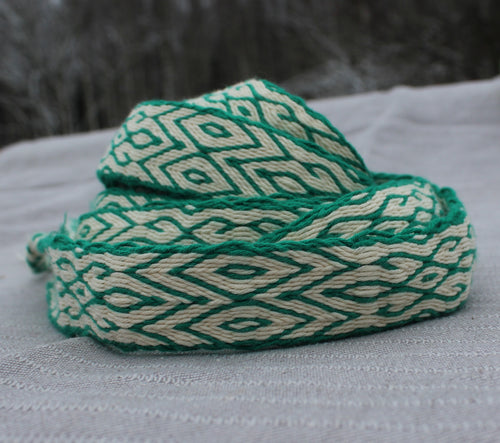 A coiled green and cream woven strap with a geometric diamond pattern, resting on a textured light gray fabric surface.