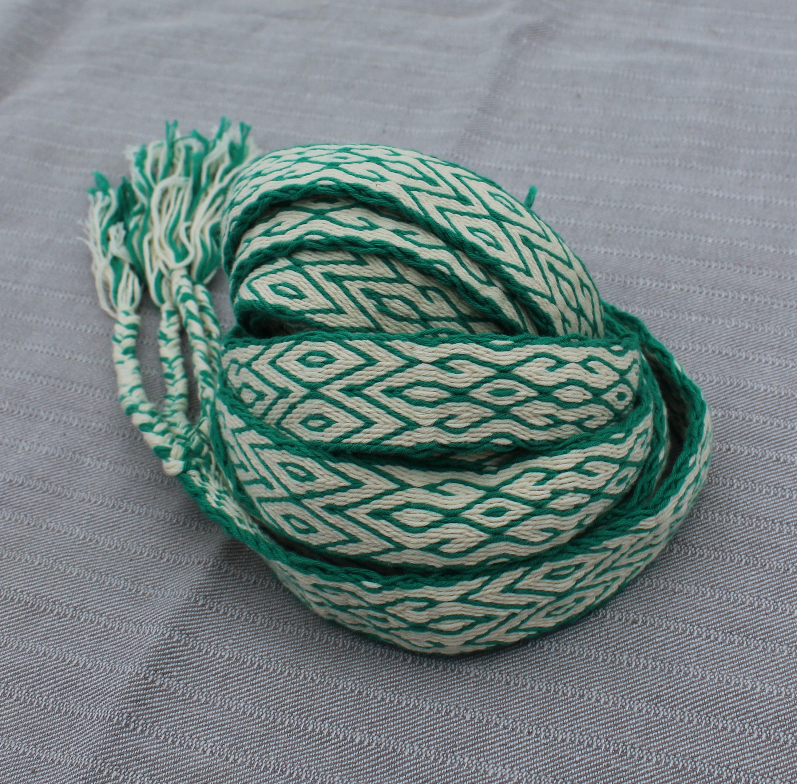 A coiled length of braided cord trim featuring a green and off-white geometric diamond pattern, with frayed ends, displayed on a textured gray fabric background.