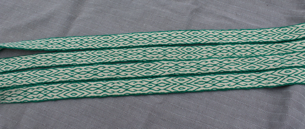 A close-up of a woven fabric strap with a repeating green and off-white geometric diamond pattern, laid flat on a textured gray background.