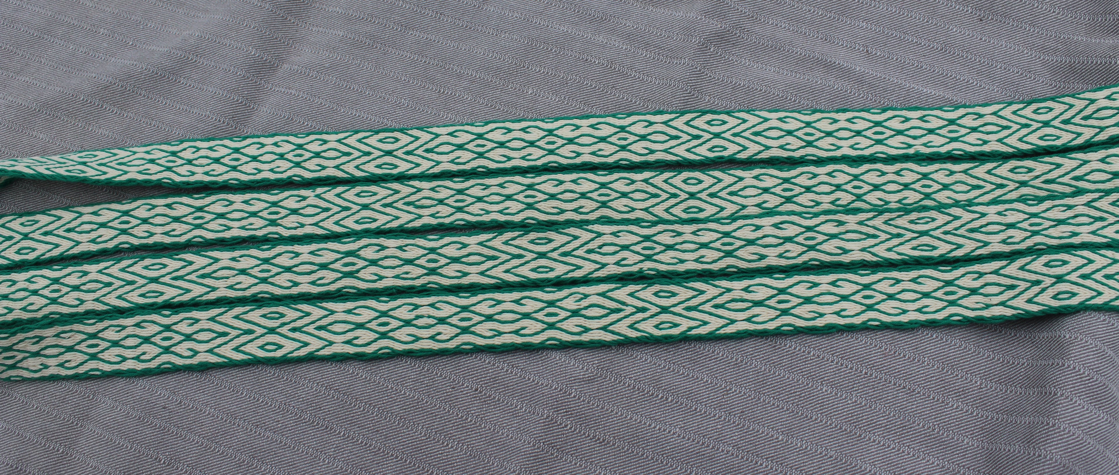 A close-up of a woven fabric strap with a repeating green and off-white geometric diamond pattern, laid flat on a textured gray background.