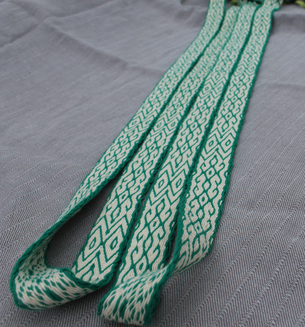 A close-up of a woven strap or belt with a green and off-white geometric pattern, laid diagonally on a textured gray fabric background.