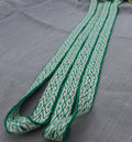A close-up of a woven strap or belt with a green and off-white geometric pattern, laid diagonally on a textured gray fabric background.