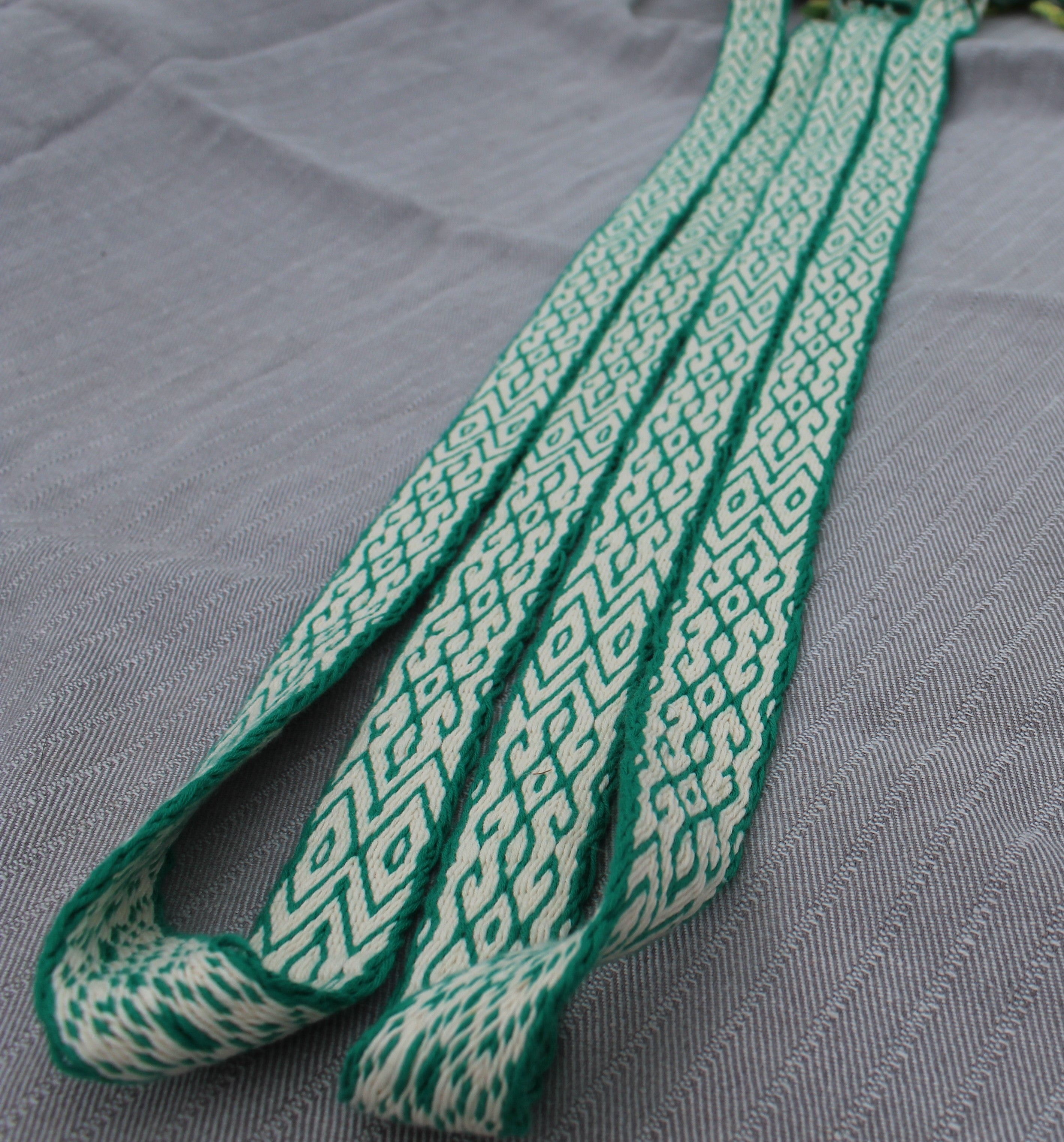 A close-up of a woven strap or belt with a green and off-white geometric pattern, laid diagonally on a textured gray fabric background.