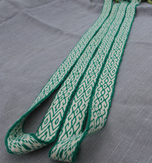 A close-up of a woven strap or belt with a green and off-white geometric pattern, laid diagonally on a textured gray fabric background.