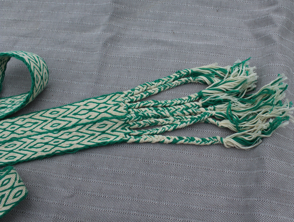 A close-up of a woven textile accessory, likely a belt or strap, featuring a green and off-white geometric diamond pattern with frayed tassels, laid on a textured gray fabric background.