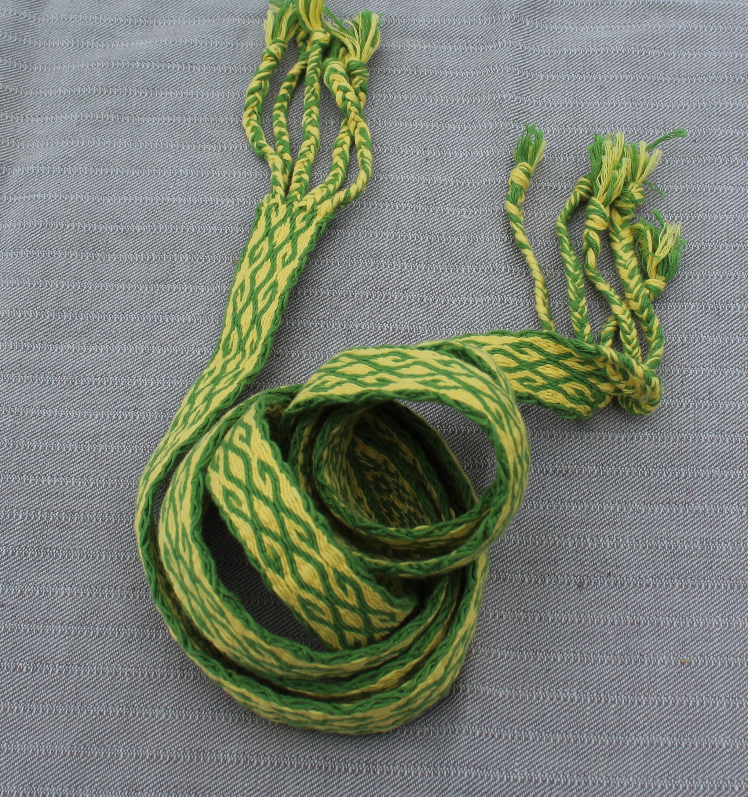 A green and yellow patterned braided strap or cord with frayed ends, coiled on a textured gray background.