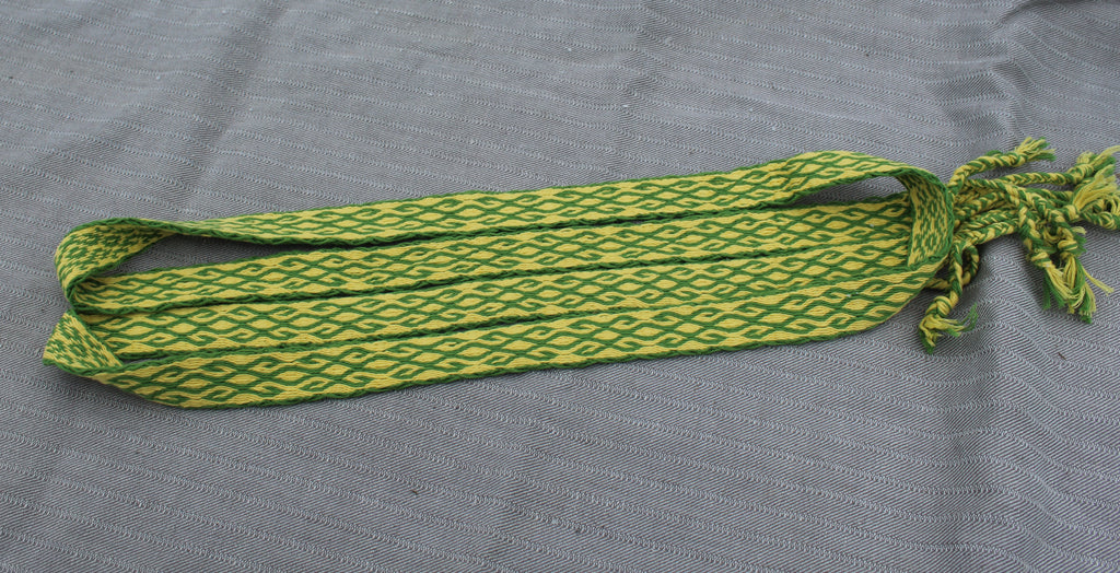 A woven waist belt with a green and yellow geometric pattern, laid flat on a gray textured fabric background.