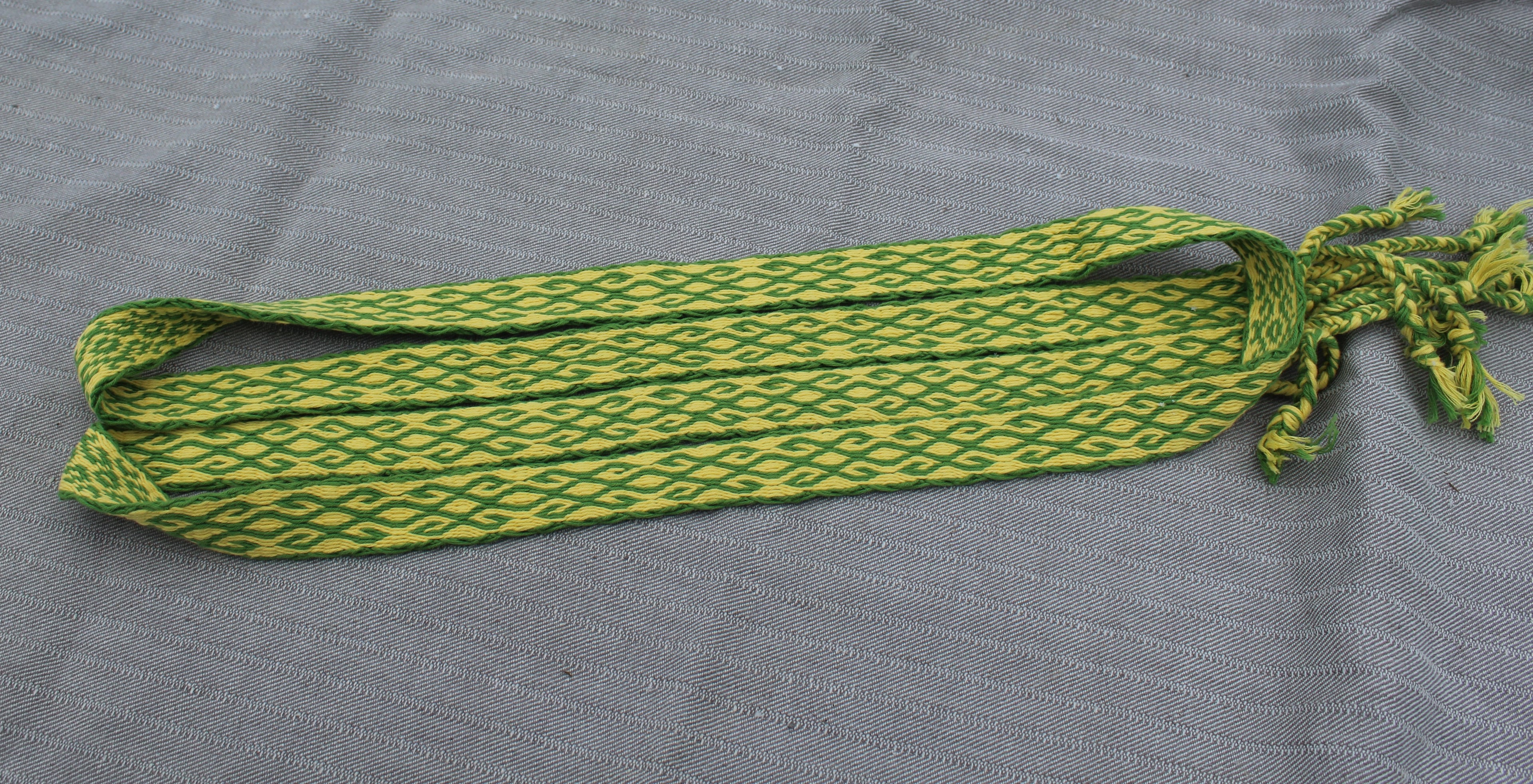 A woven waist belt with a green and yellow geometric pattern, laid flat on a gray textured fabric background.