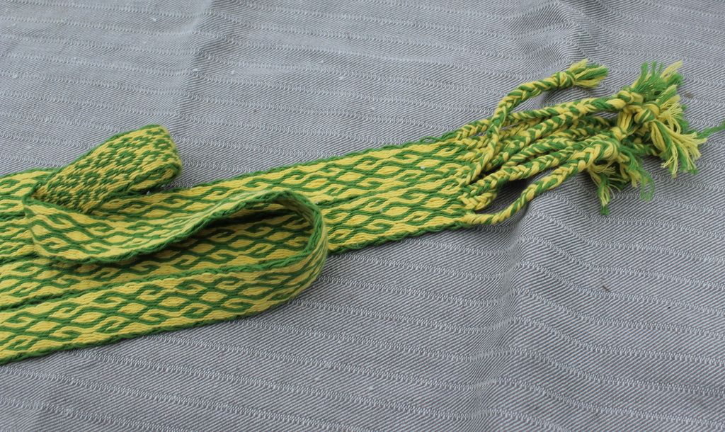 A green and yellow patterned woven strap bag with knotted tassels, laid flat on a textured gray fabric background.